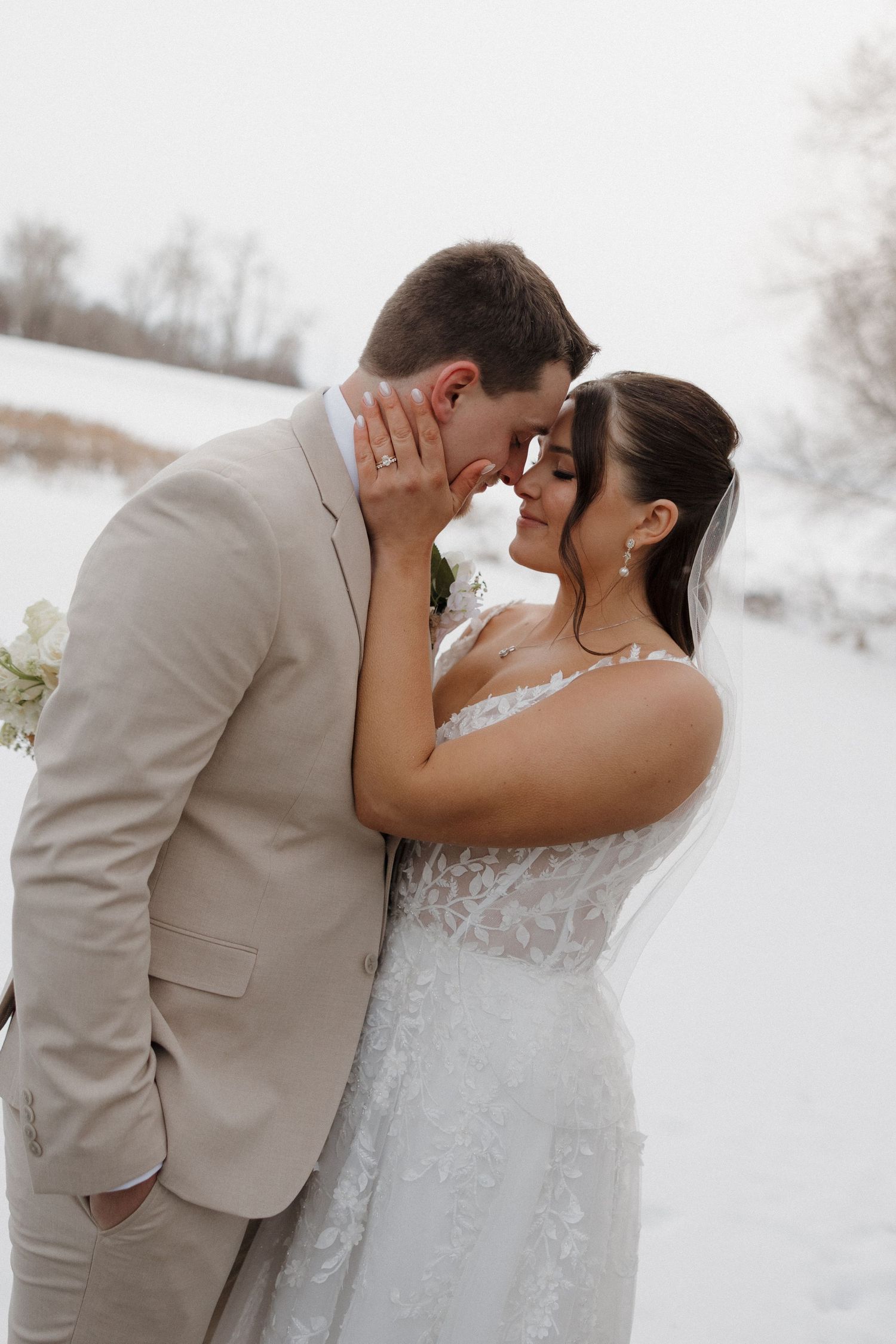 A tender moment between newlyweds in a snowy winter landscape, with the bride in lace and groom in beige suit.