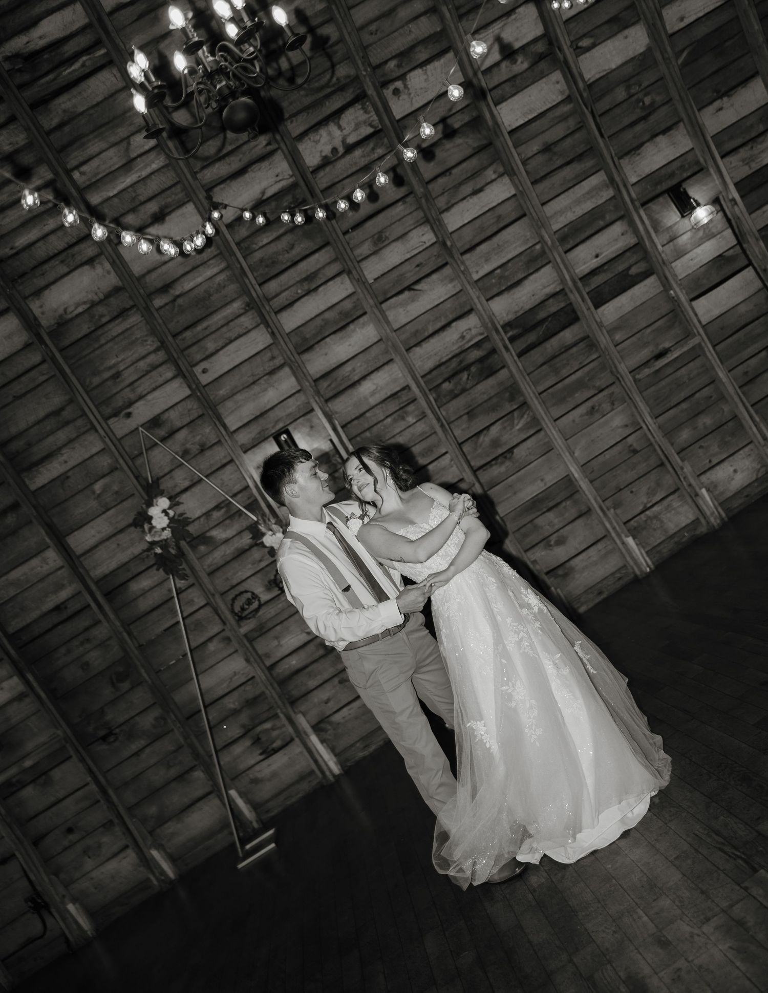 A couple dances intimately on a rustic wooden floor under vintage string lights in black and white photography.