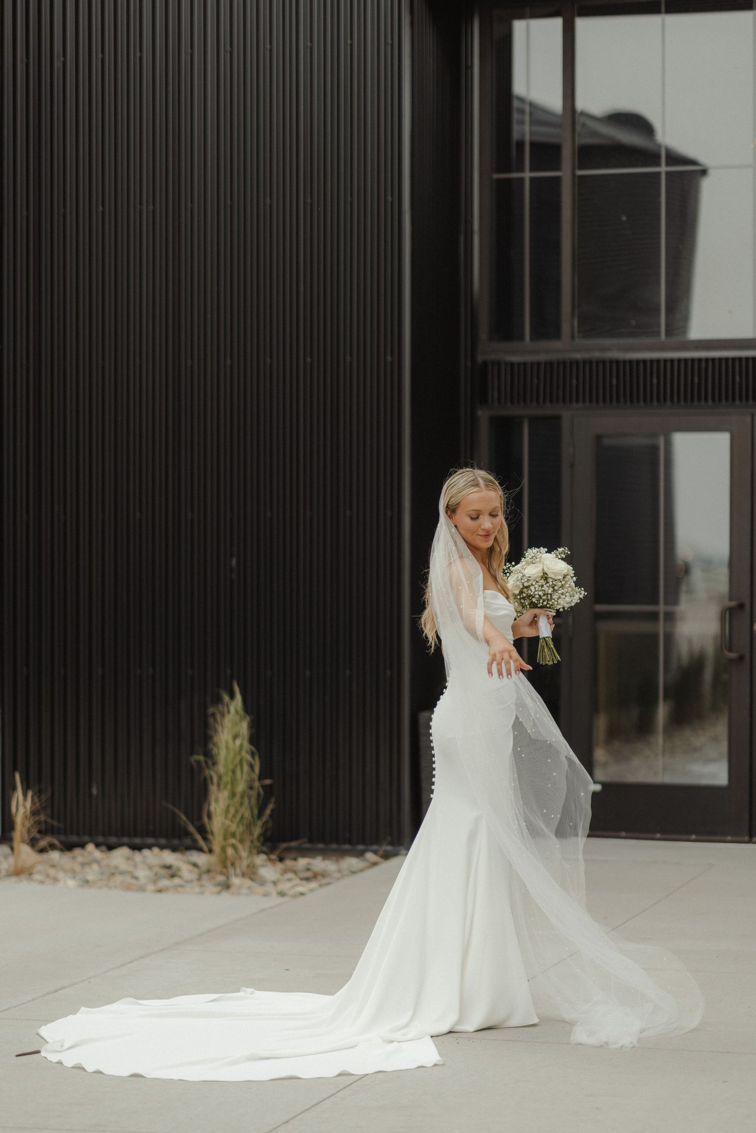 A bride in a flowing white gown with a long train poses elegantly against a modern black building exterior.
