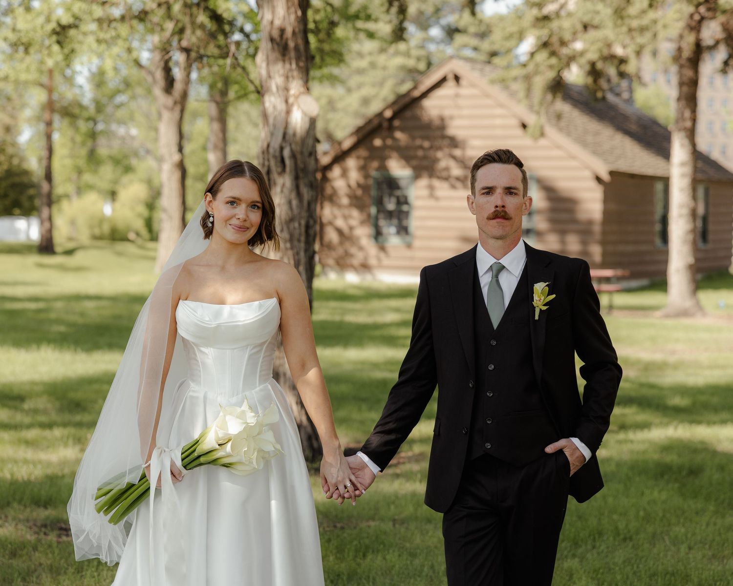 A couple in formal wedding attire holds hands while walking through a sunlit wooded area with a rustic cabin in background.