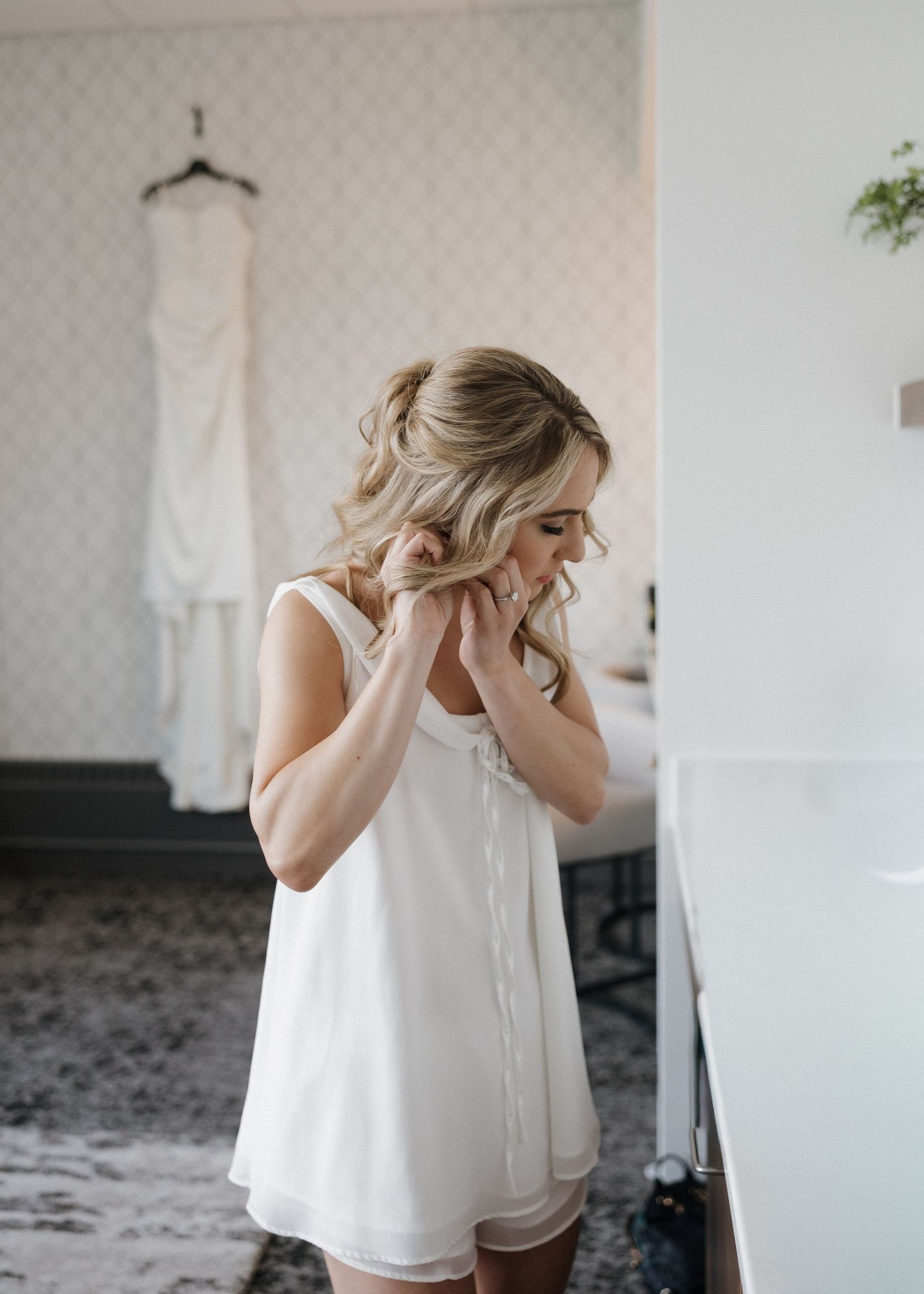 Bride in white slip dress putting on earrings before wedding ceremony with gown hanging in background.