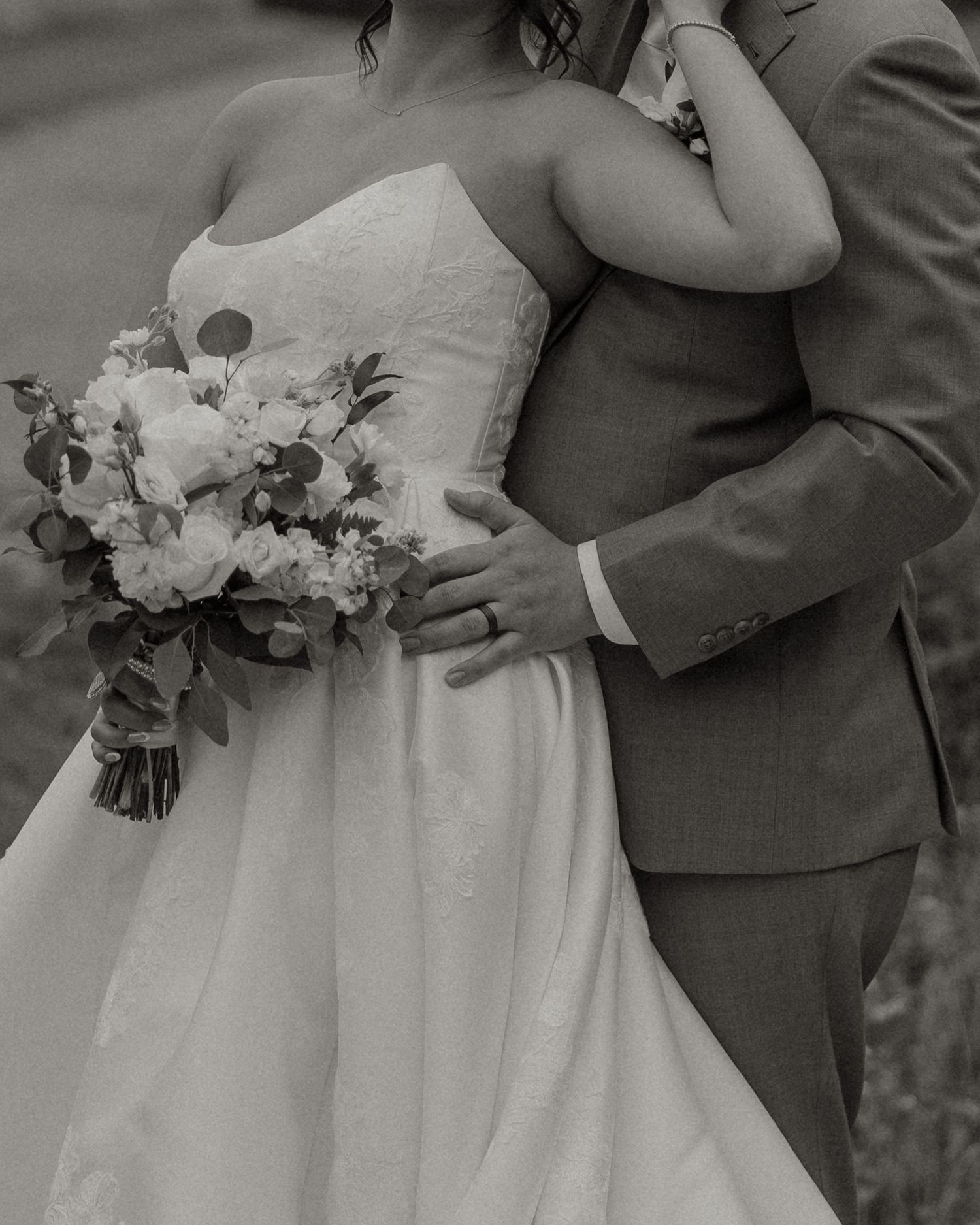 Black and white photo of bride in strapless gown holding floral bouquet embracing groom in gray suit.