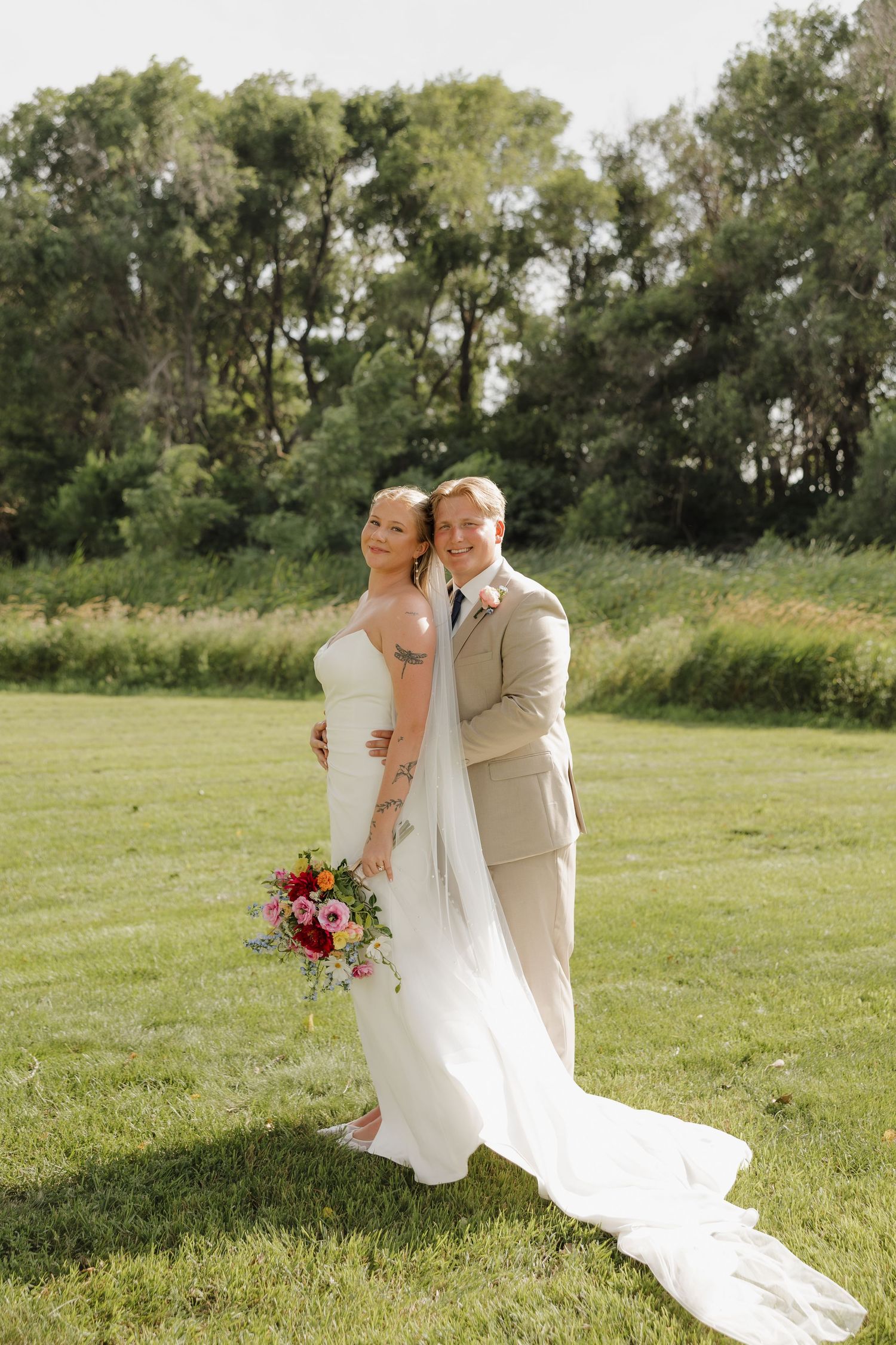 Newlywed couple embraces on a sunny day in a lush green field during their outdoor wedding photoshoot.