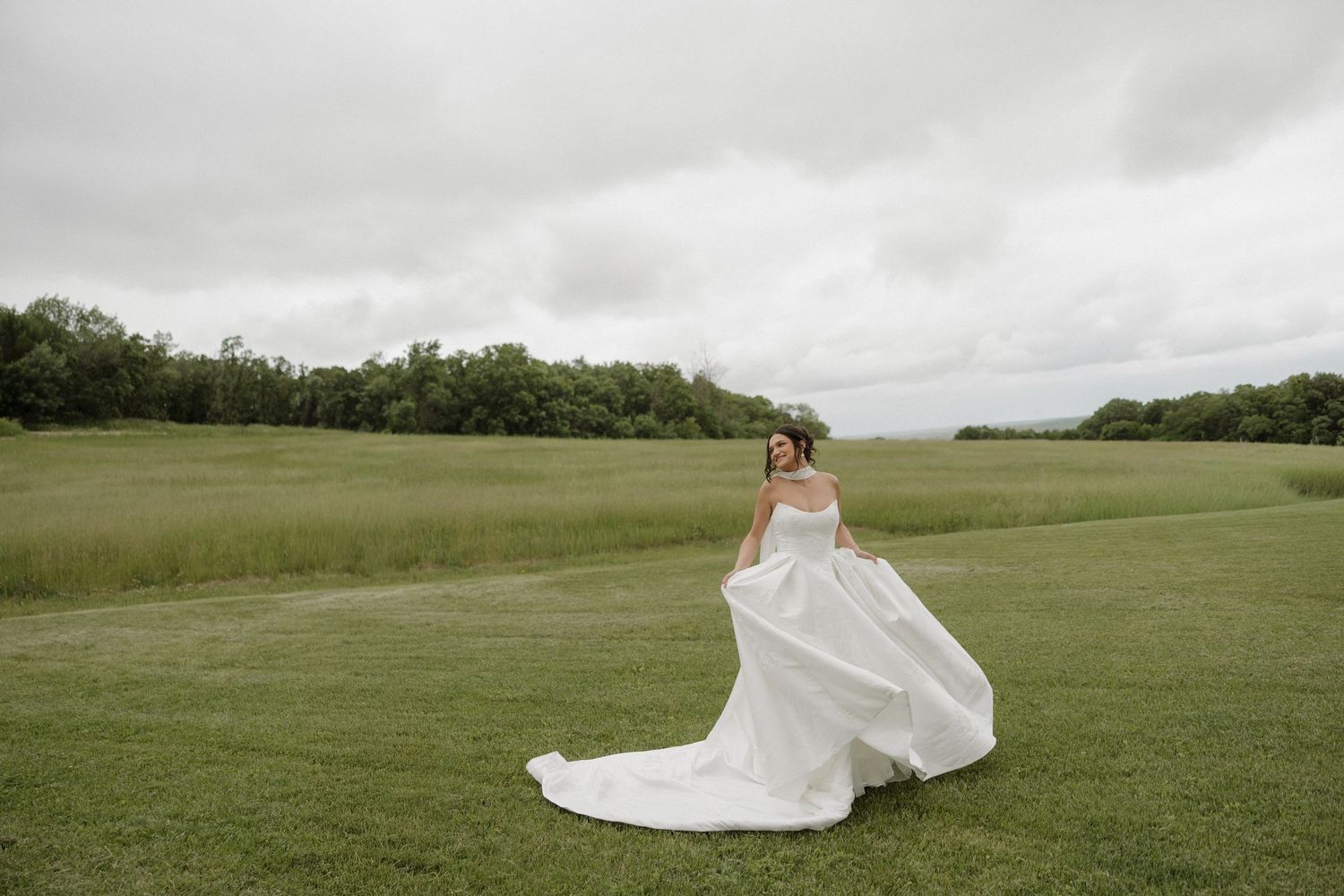 Bride in a flowing white gown twirling in a lush green field with trees and cloudy sky backdrop.
