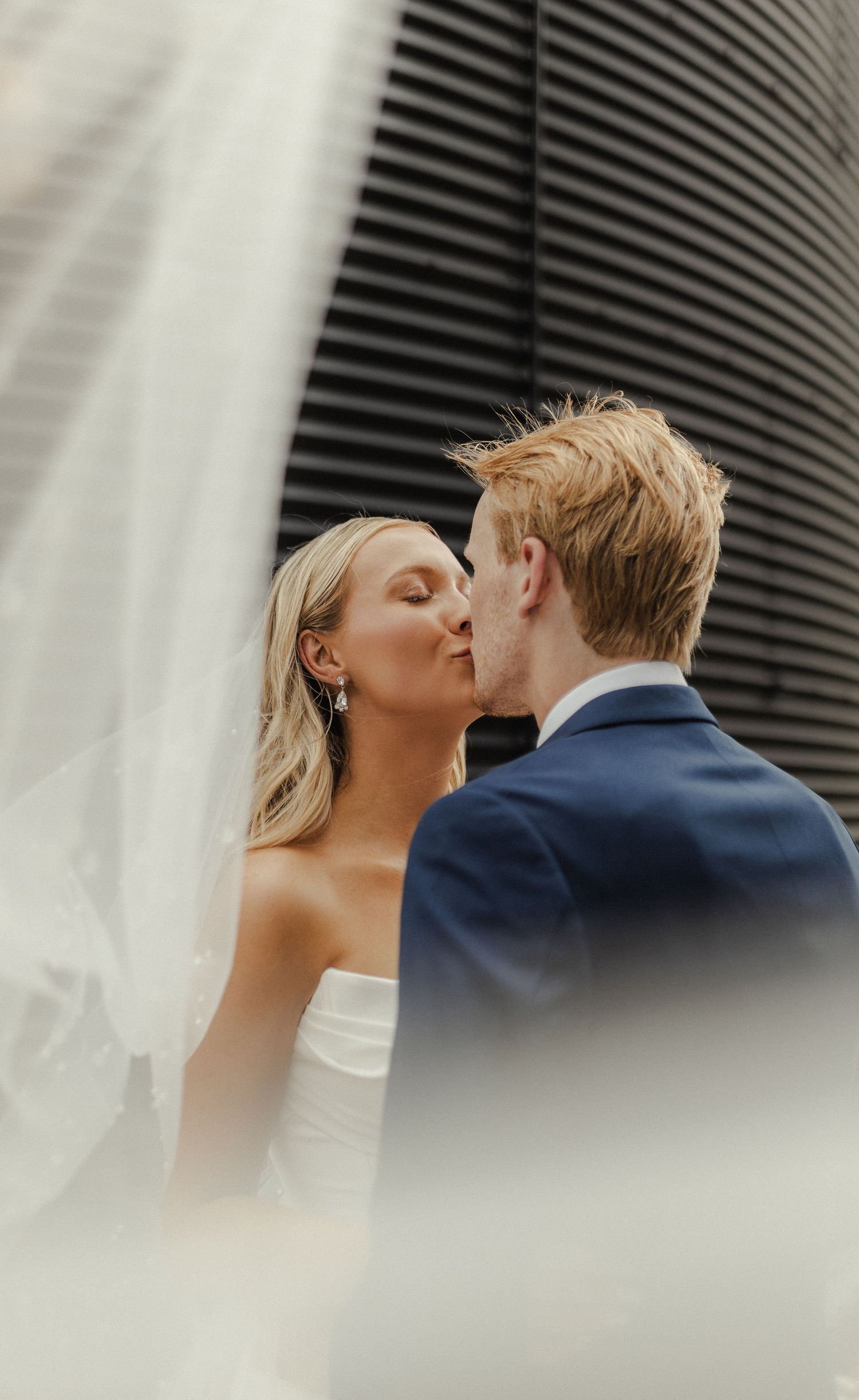 A bride and groom share a romantic kiss during their wedding photoshoot against a modern architectural backdrop.