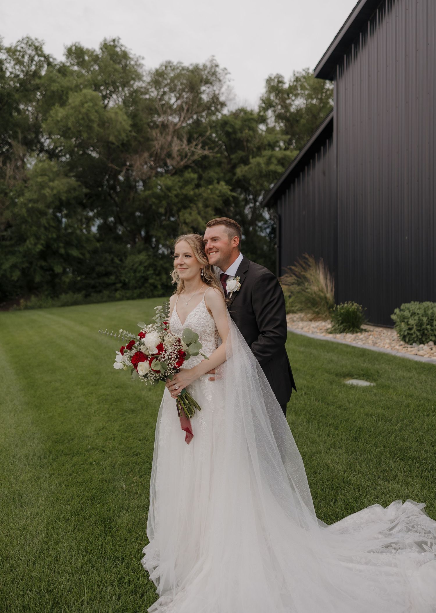 Newlywed couple shares romantic moments on their wedding day against black barn and lush green landscape.