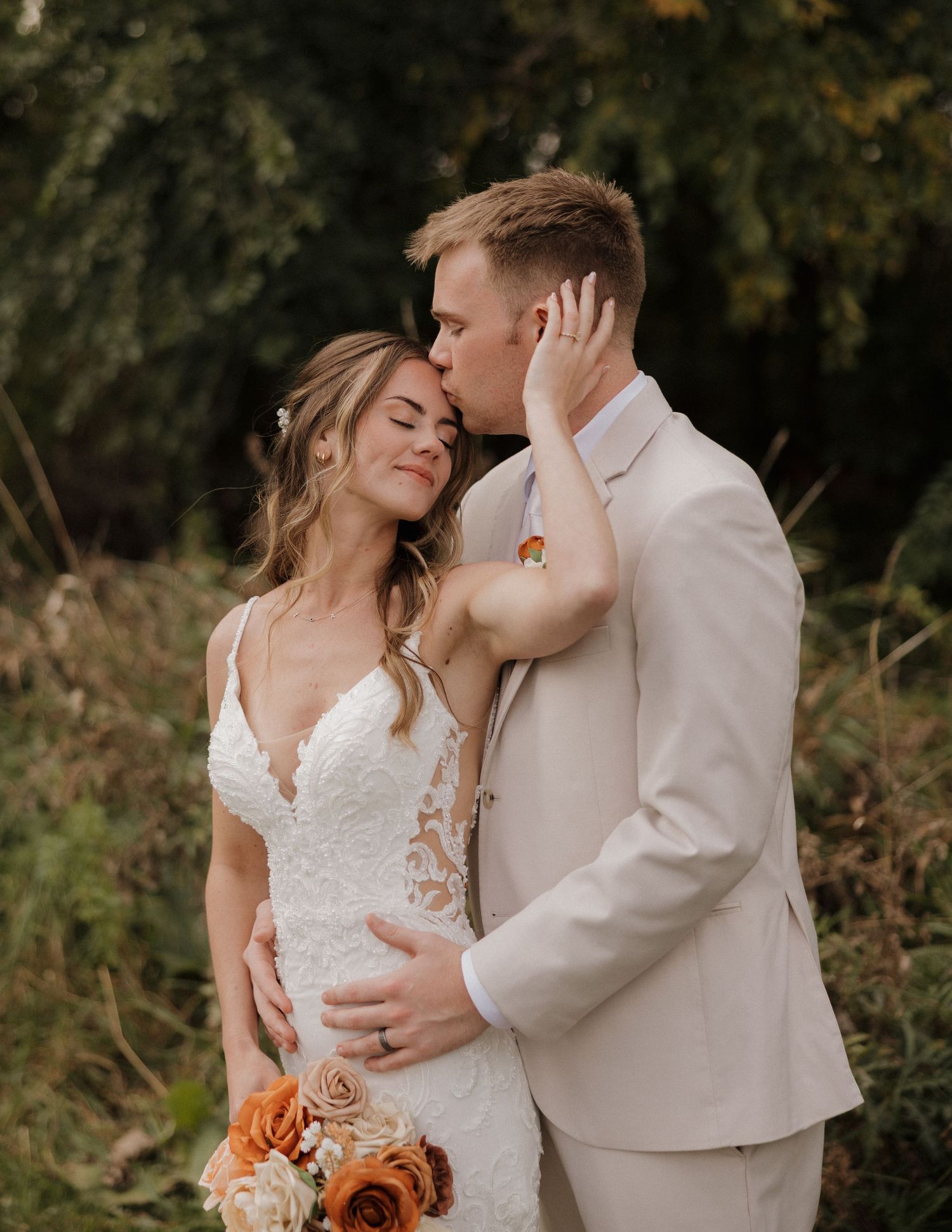 A romantic wedding couple embraces outdoors with autumn foliage in the background and orange roses in the bouquet.