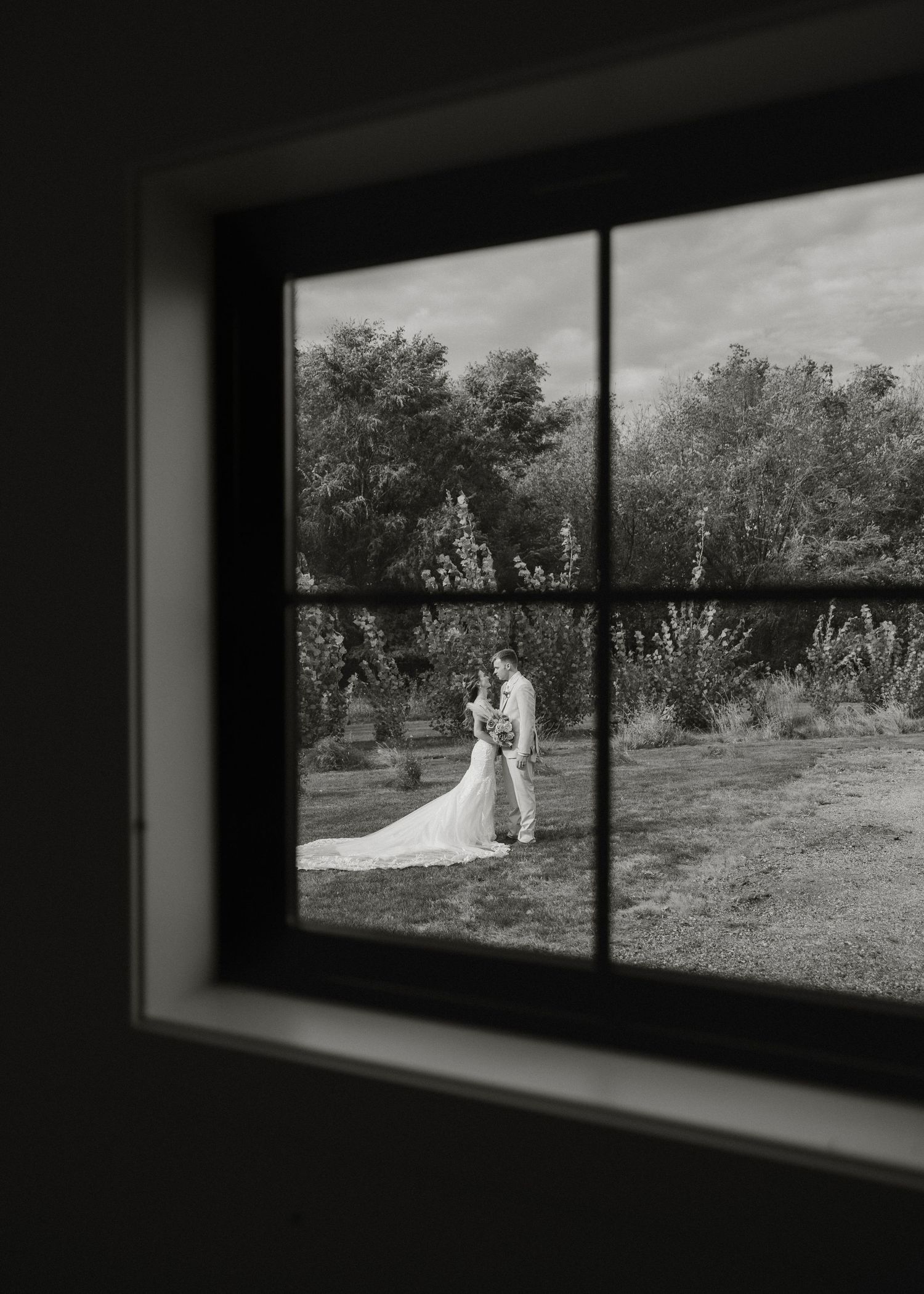 Black and white wedding photo framed through a window shows a couple embracing in a garden setting.