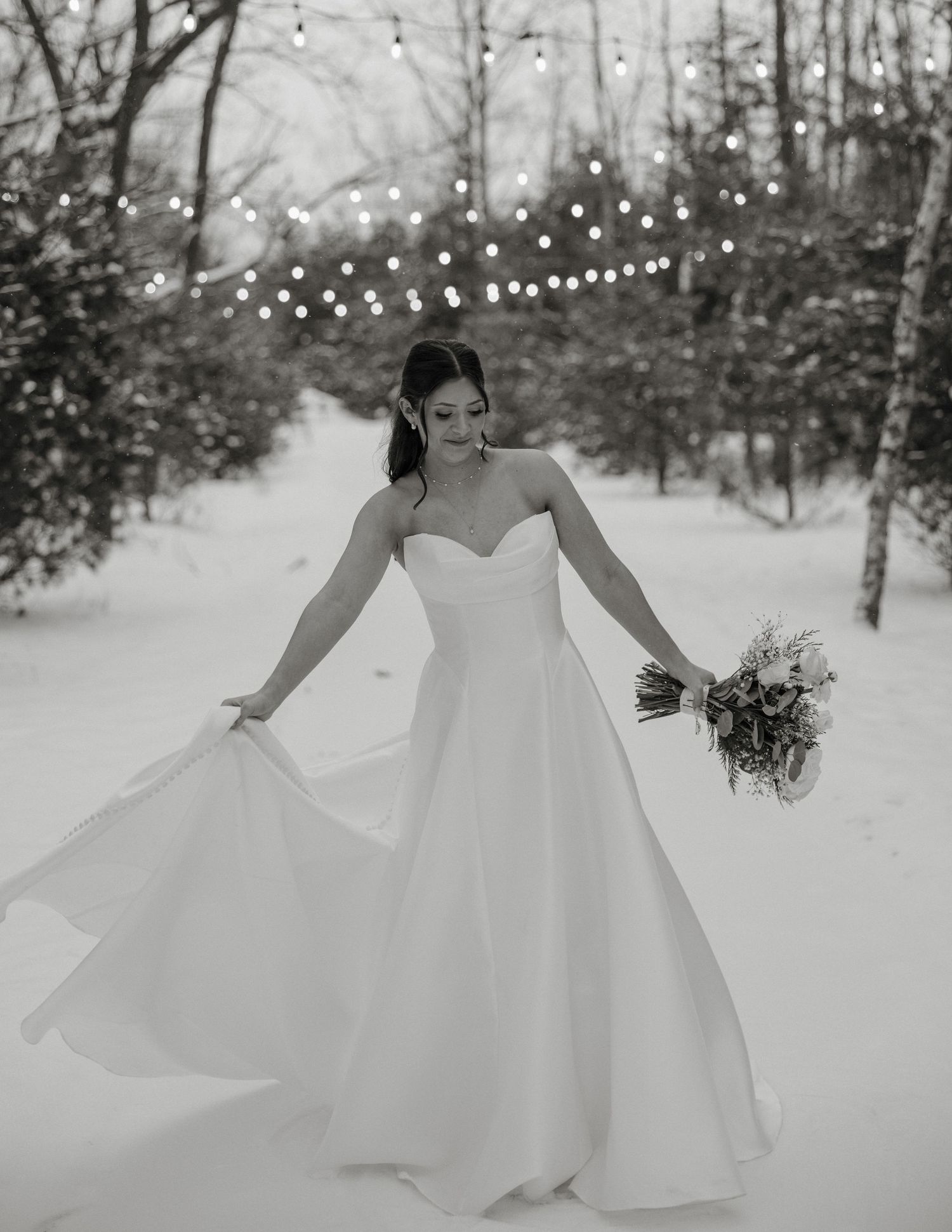 A bride in a flowing white gown twirls gracefully in a snowy winter forest setting with string lights overhead.