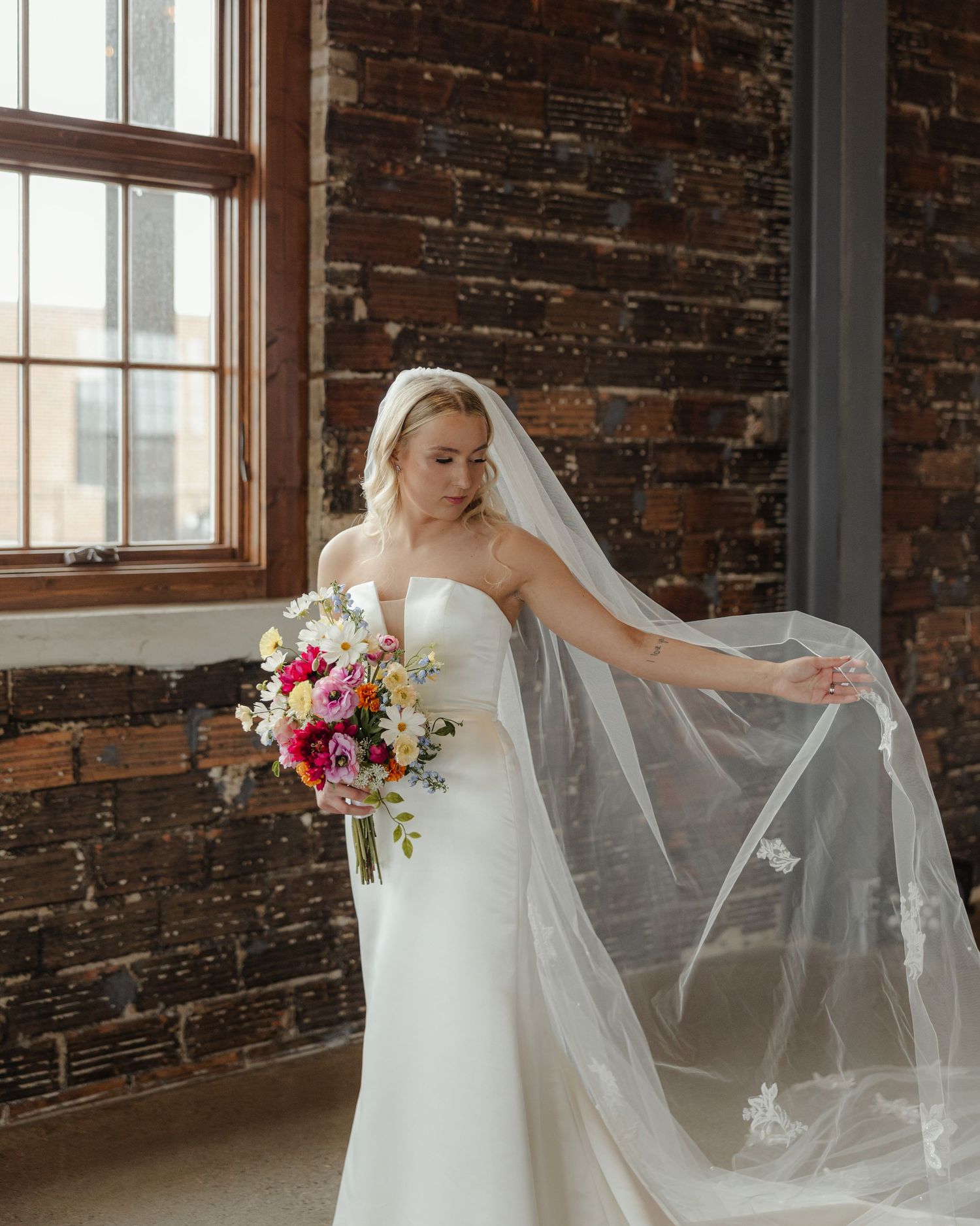 A bride in a white strapless gown holds a vibrant pink and red bouquet while posing against an exposed brick wall.