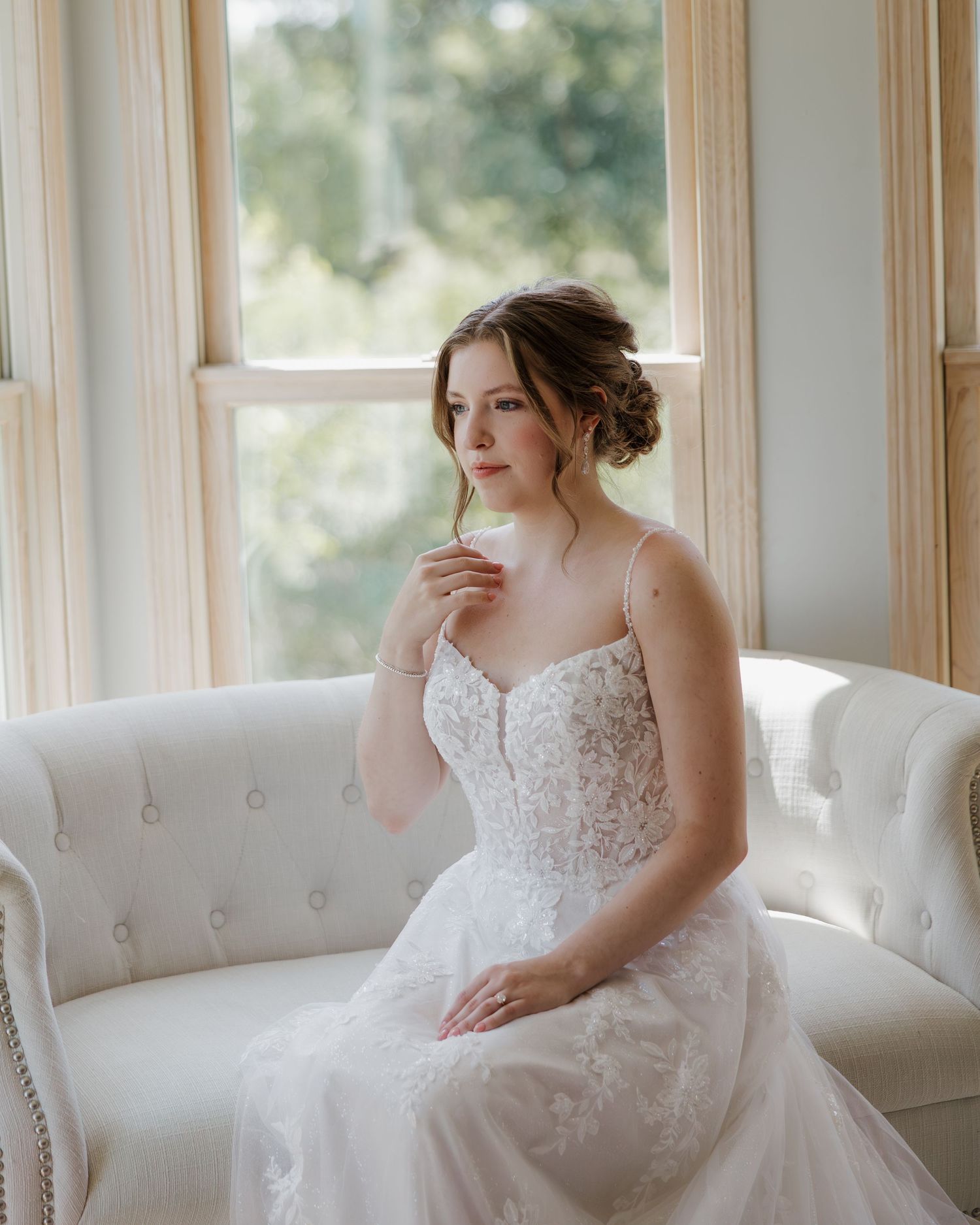 Elegant bride in lace wedding gown poses gracefully near sunlit windows in a bright white room.