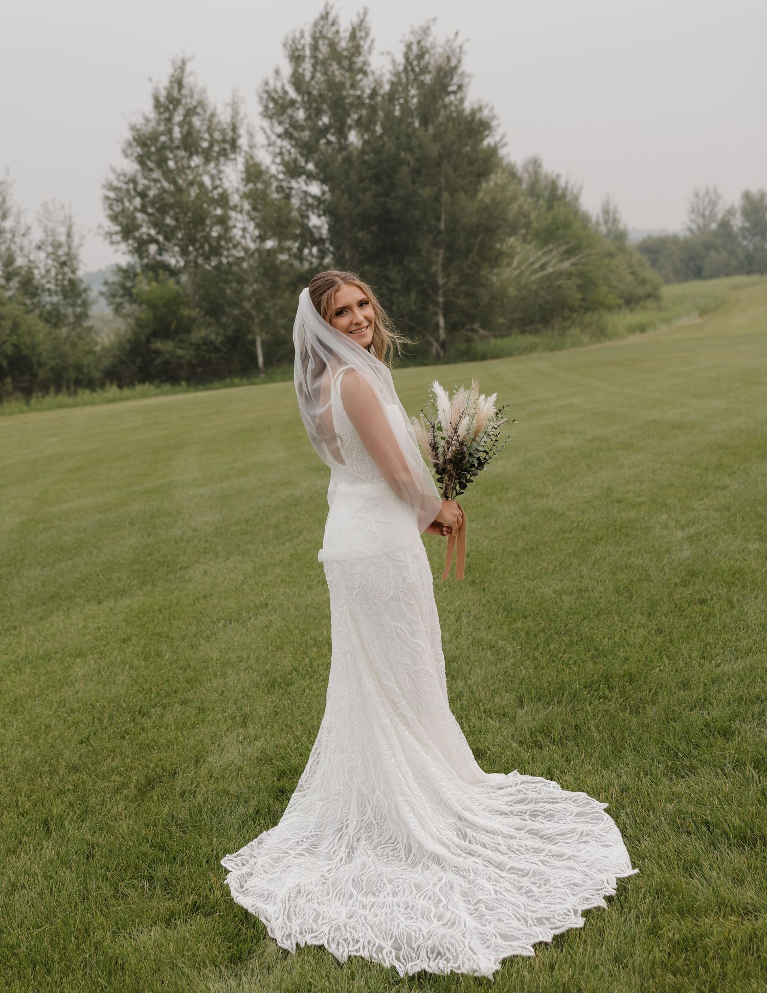 A bride in a lace wedding gown poses gracefully on a grassy hillside during an outdoor photo session.