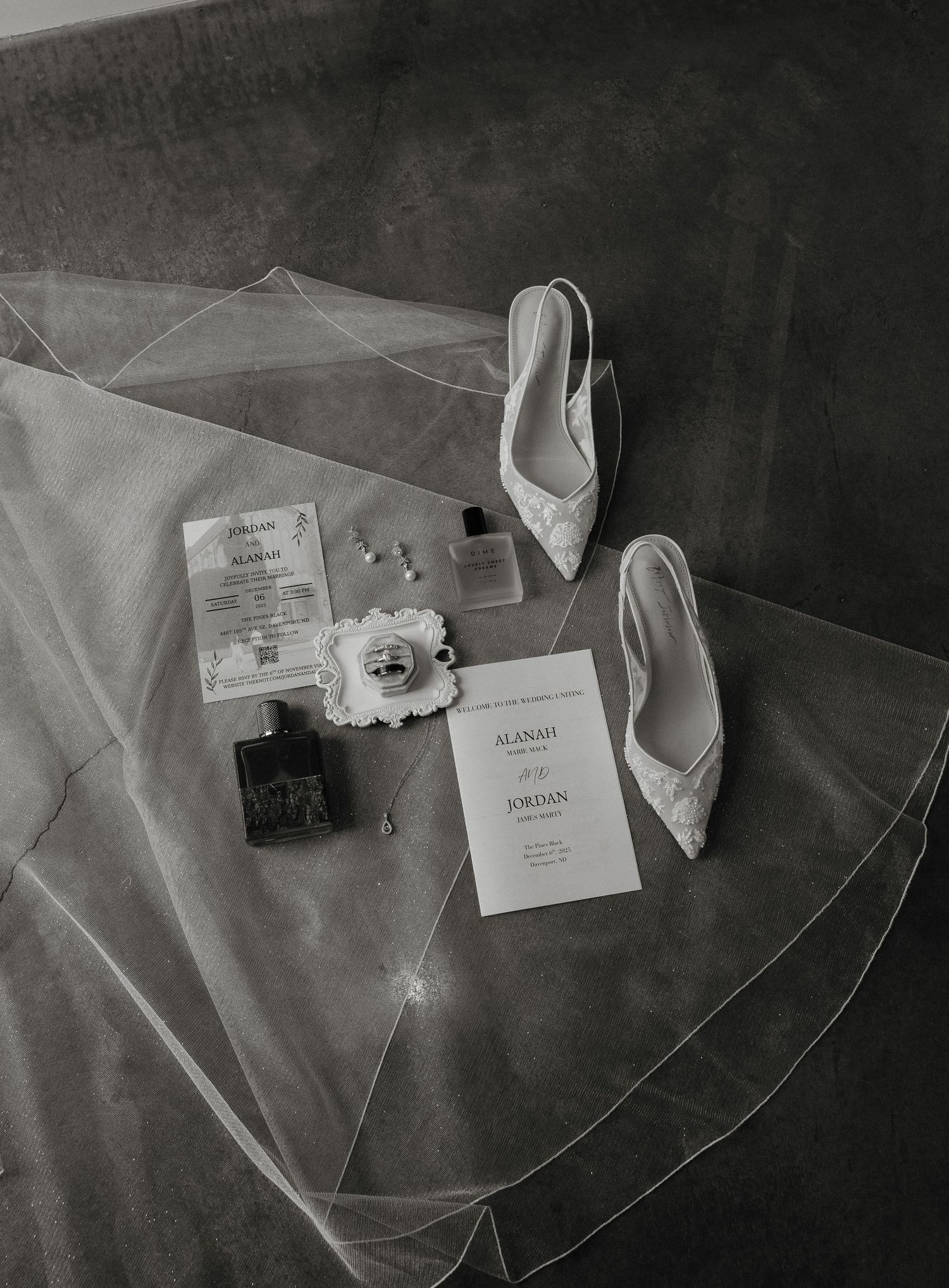 Black and white wedding details including invitation cards, white shoes, and delicate veil arranged artistically on a dark surface.