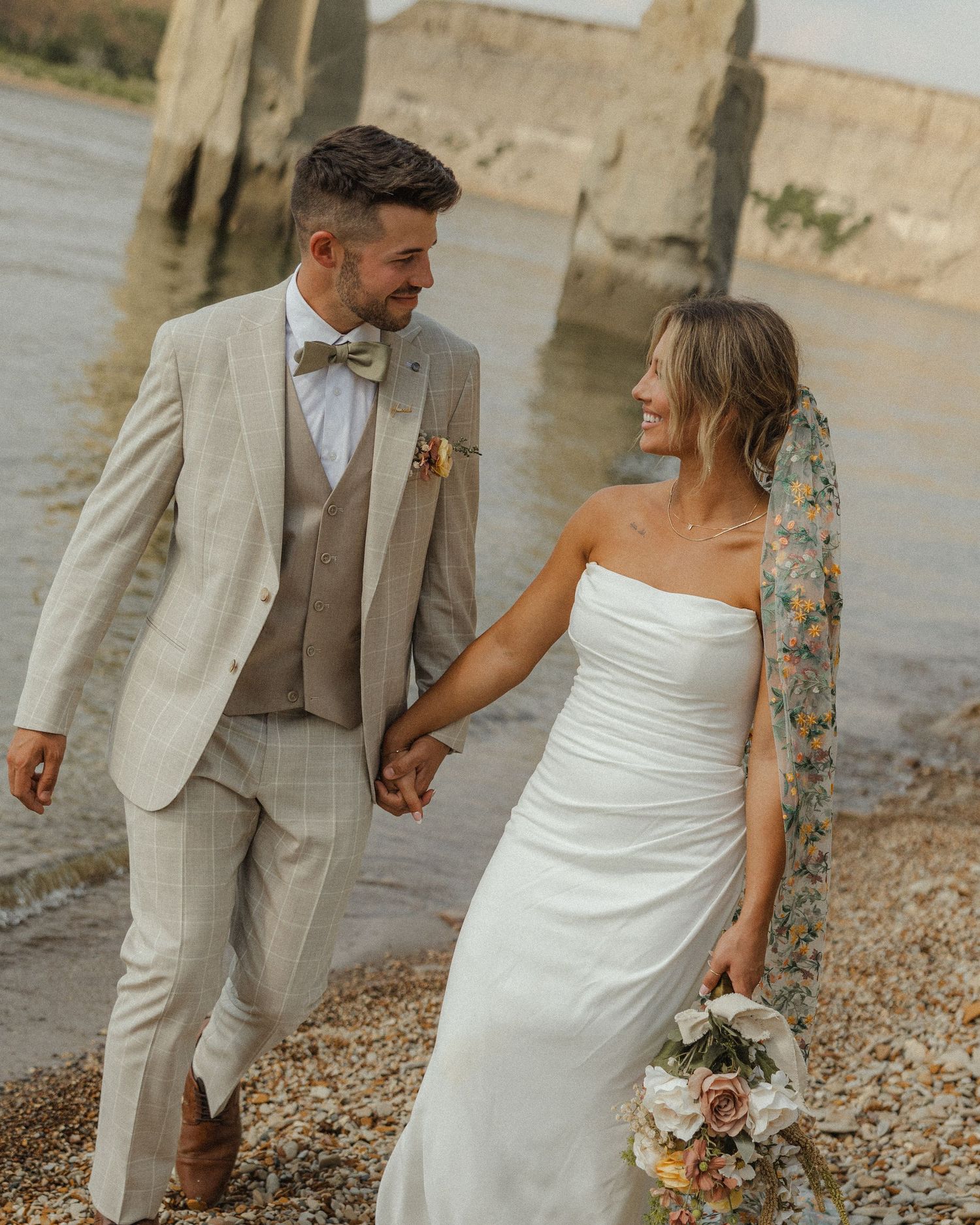 Newlyweds walking on beach, groom in beige suit and bride in fitted white dress.
