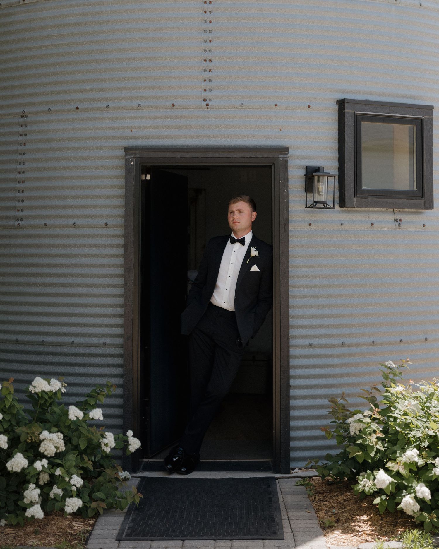 Groom in black tuxedo standing in doorway of rustic corrugated metal building surrounded by white flowers.