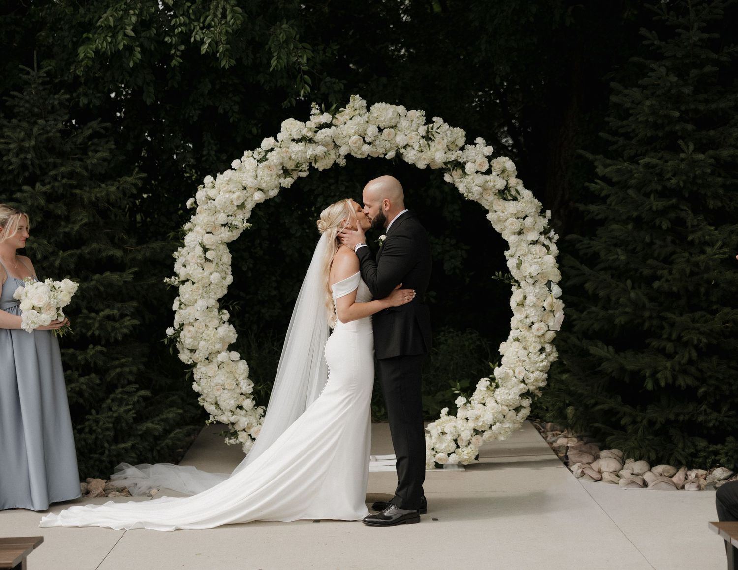 Bride and groom share first kiss under a circular white floral arch at an outdoor wedding ceremony.