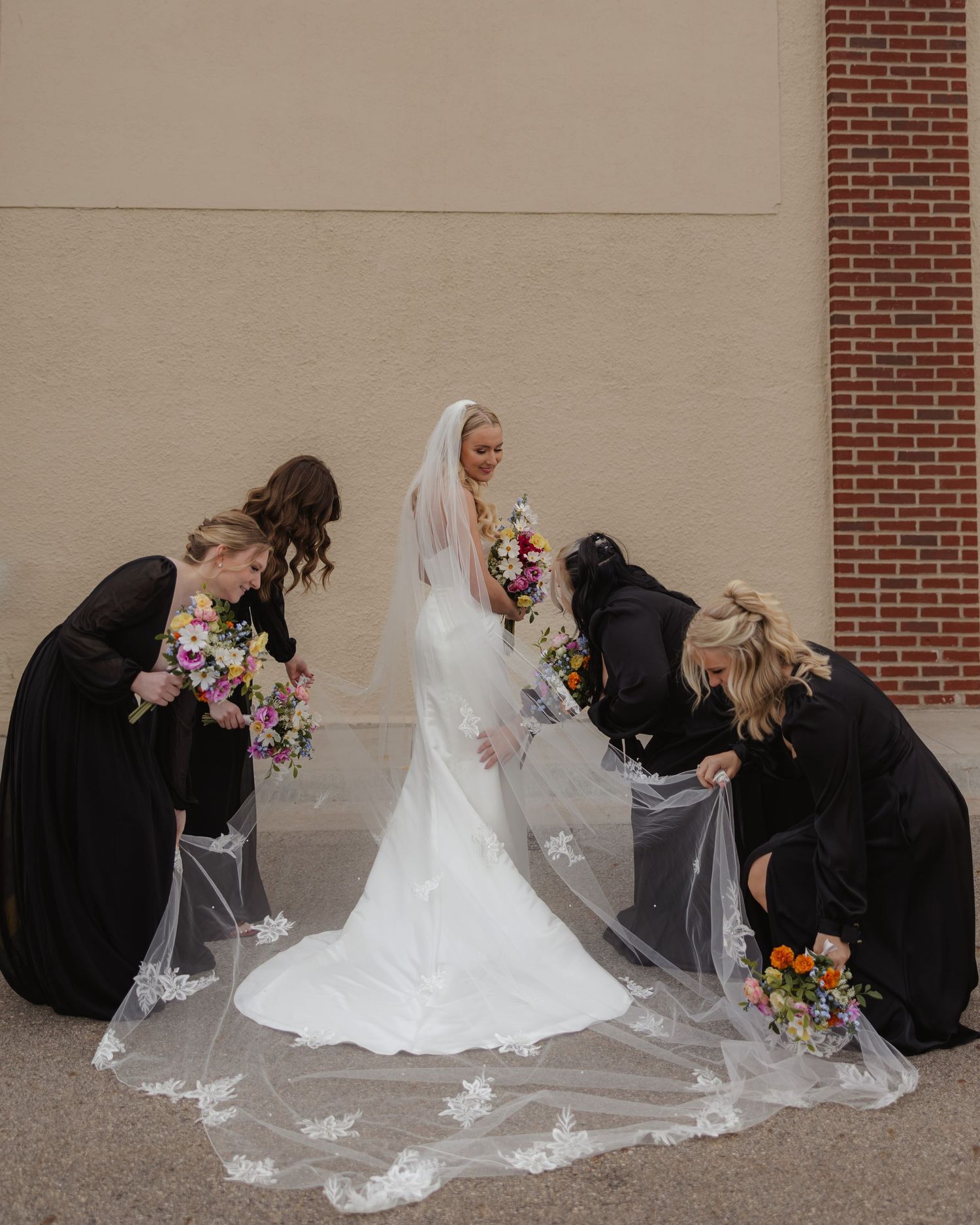 Wedding party helping arrange bride's long lace veil against brick wall background.
