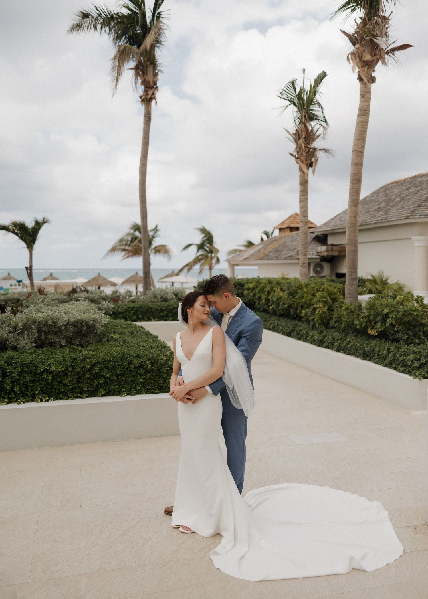 Couple in wedding attire embracing on a beachfront walkway lined with palm trees under cloudy skies.
