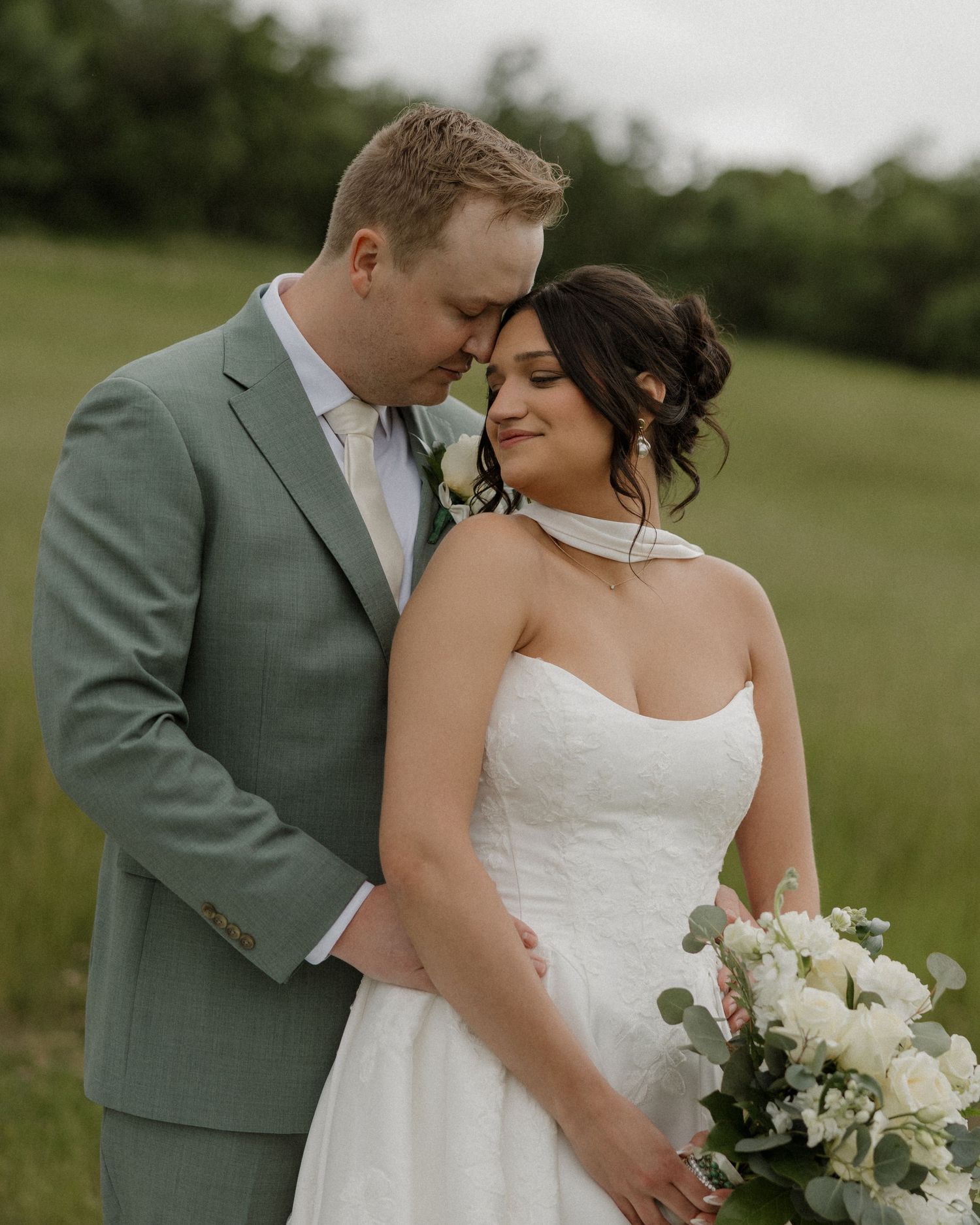 A bride in a white strapless gown holds a bouquet while a groom in a sage suit tenderly leans his head against hers outdoors.