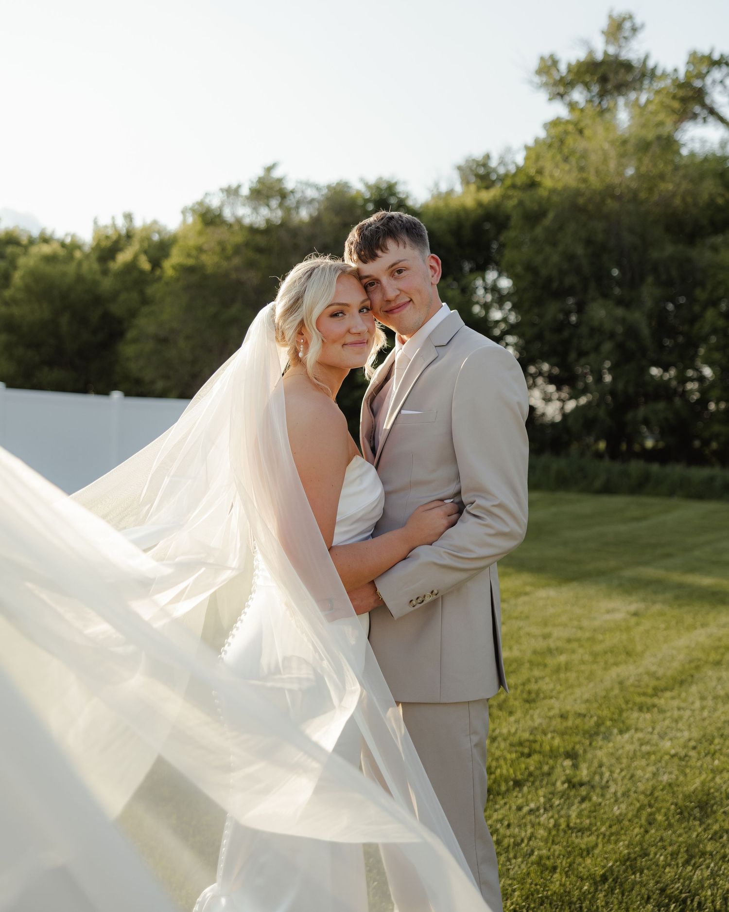 A bride in a flowing white gown and veil poses with her groom in a beige suit against a natural outdoor backdrop.