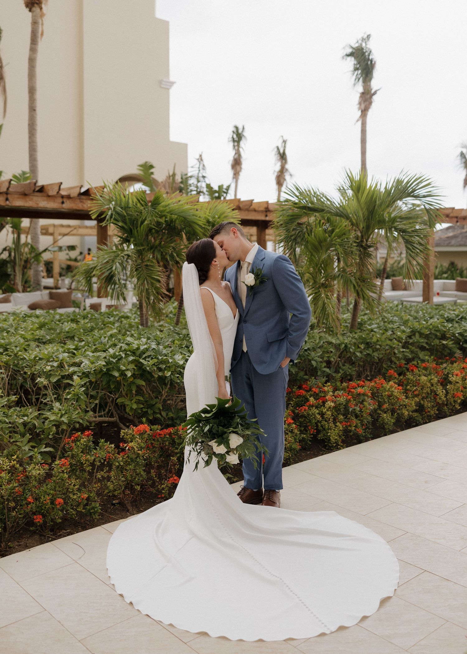 A couple shares a romantic moment in their wedding attire on a palm-lined walkway at a tropical resort.