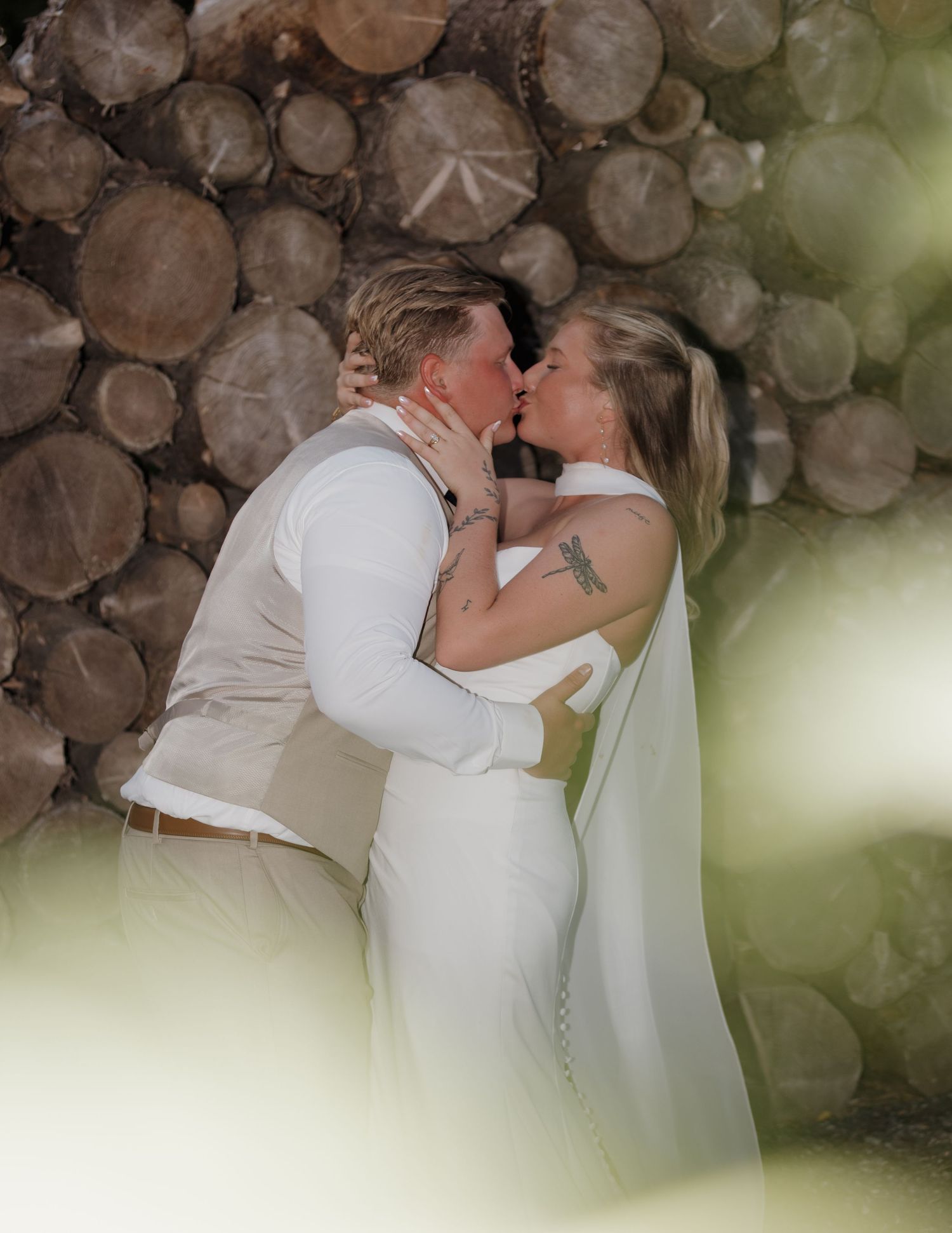 A couple in wedding attire sharing a romantic kiss against a rustic backdrop of stacked logs.