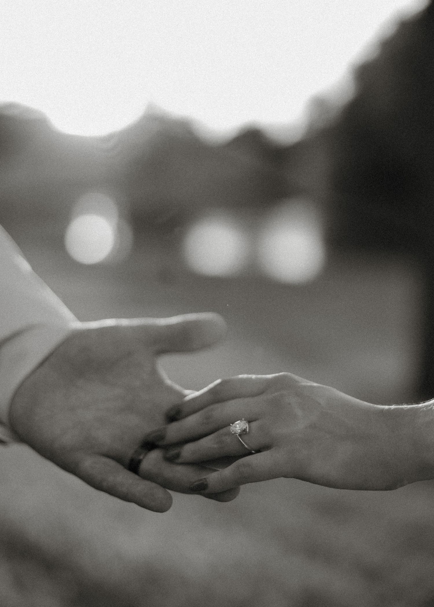A black and white close-up photo of two hands reaching for each other with an engagement ring visible.