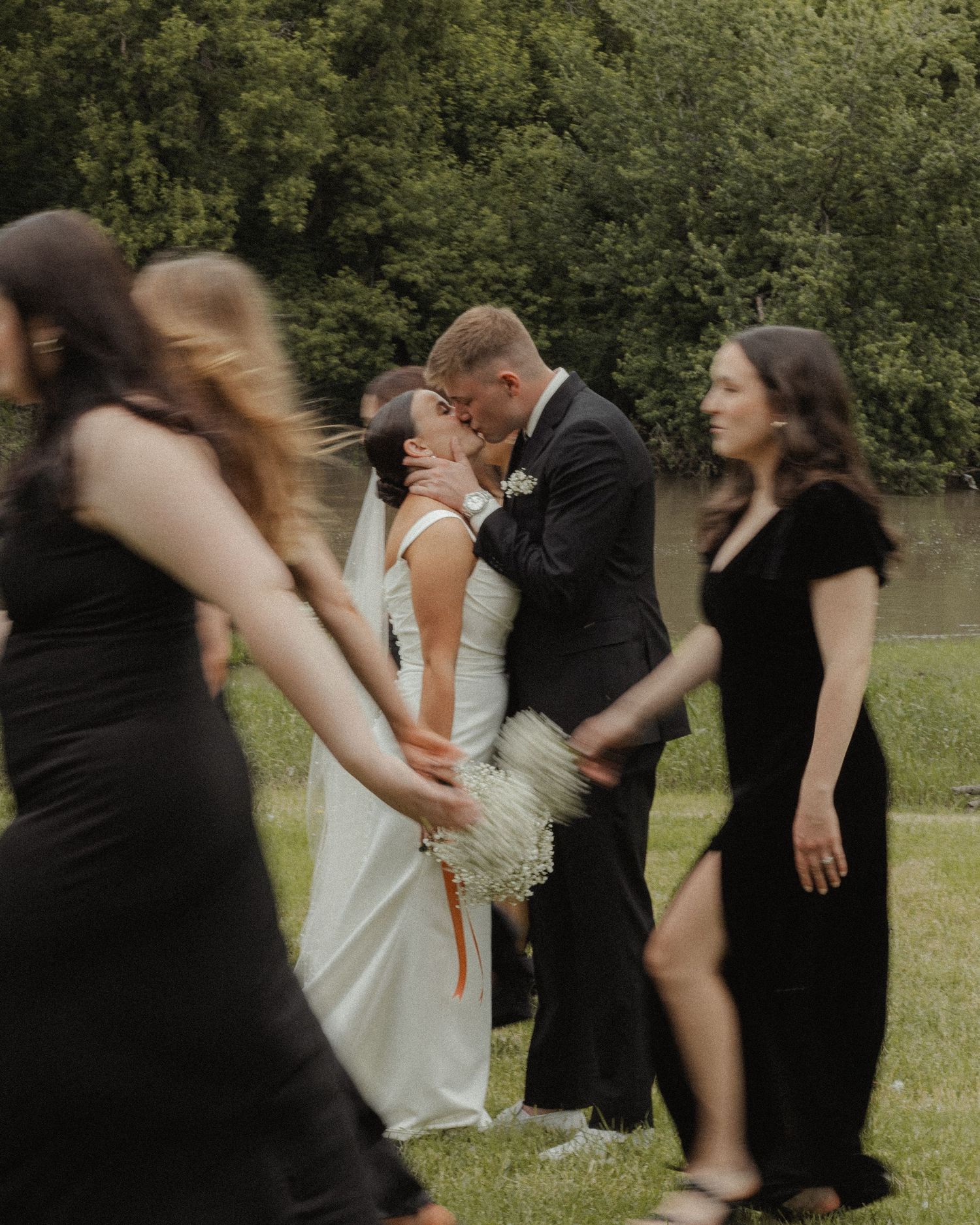 A couple shares a romantic kiss during an outdoor wedding ceremony while attendees in black dresses look on.