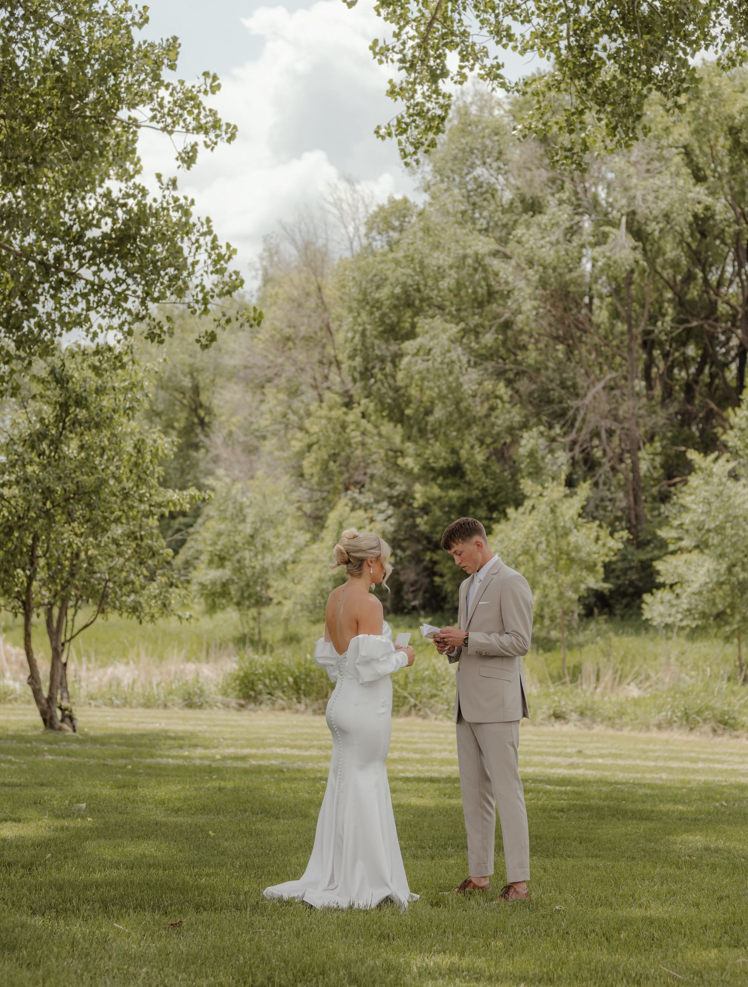 A couple shares an intimate moment in a lush green meadow surrounded by trees on their wedding day.
