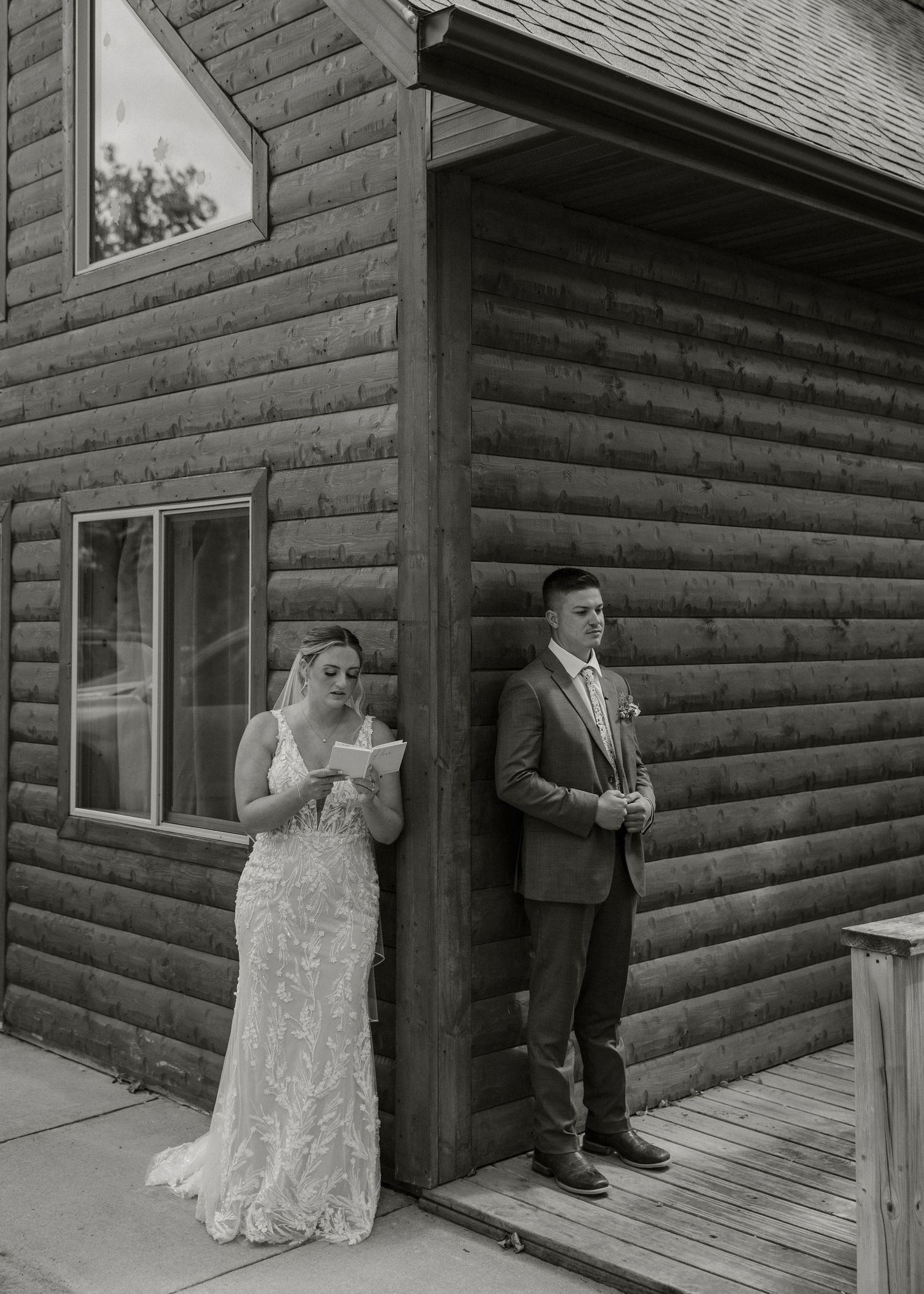 A black and white photo of a couple in wedding attire standing outside a rustic log cabin on a wooden deck.