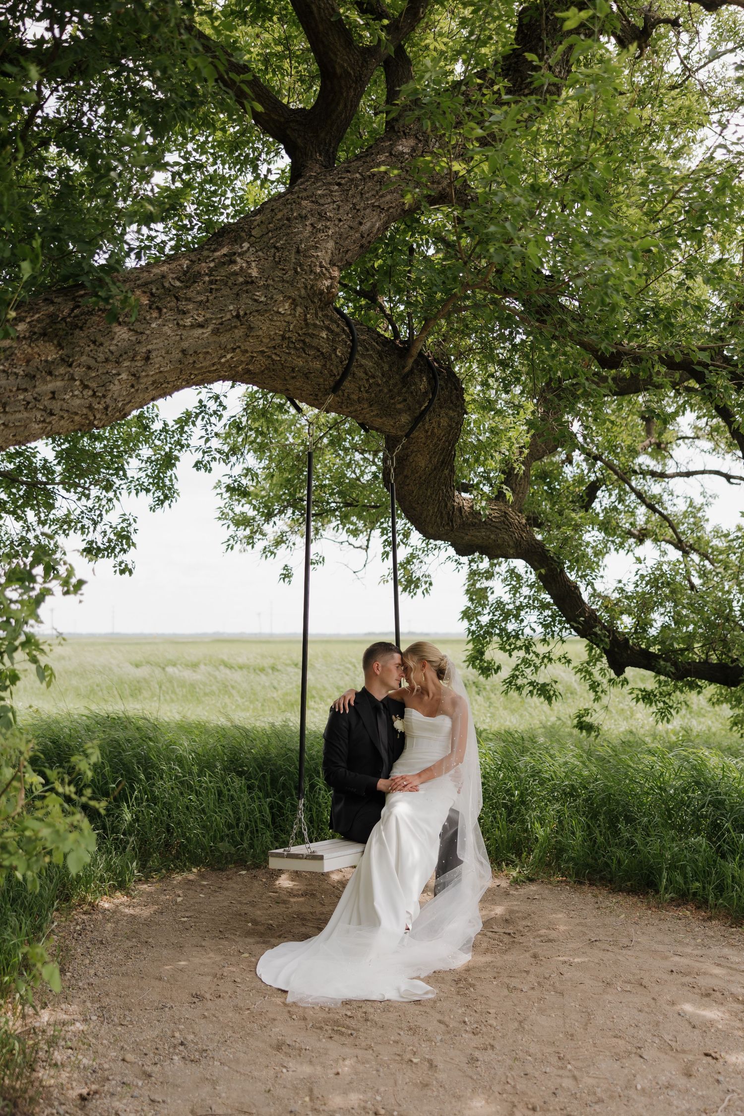 A couple shares an intimate moment under a large tree on their wedding day in a natural outdoor setting.