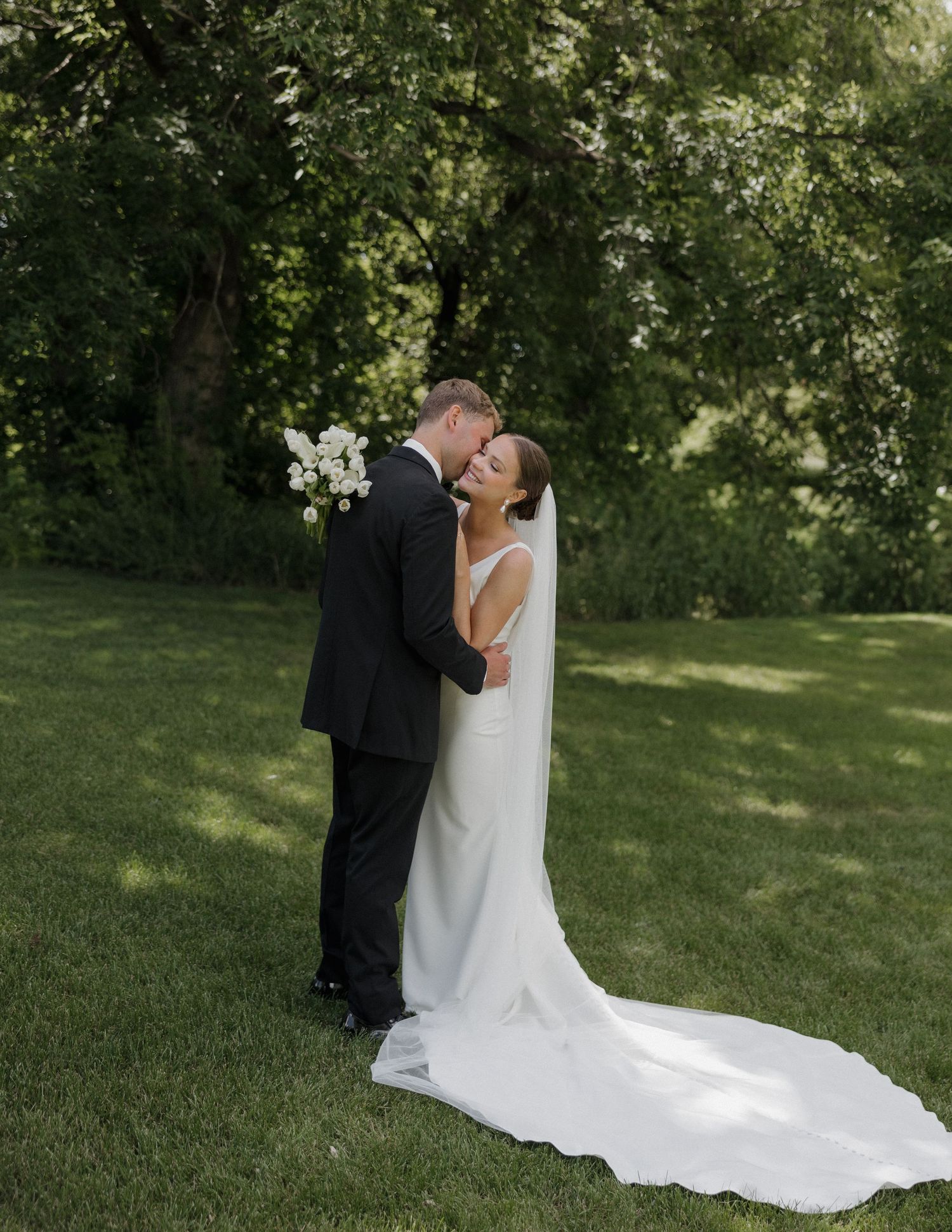 A bride and groom share a romantic embrace on a lush green lawn, with the bride in a flowing white gown and veil.