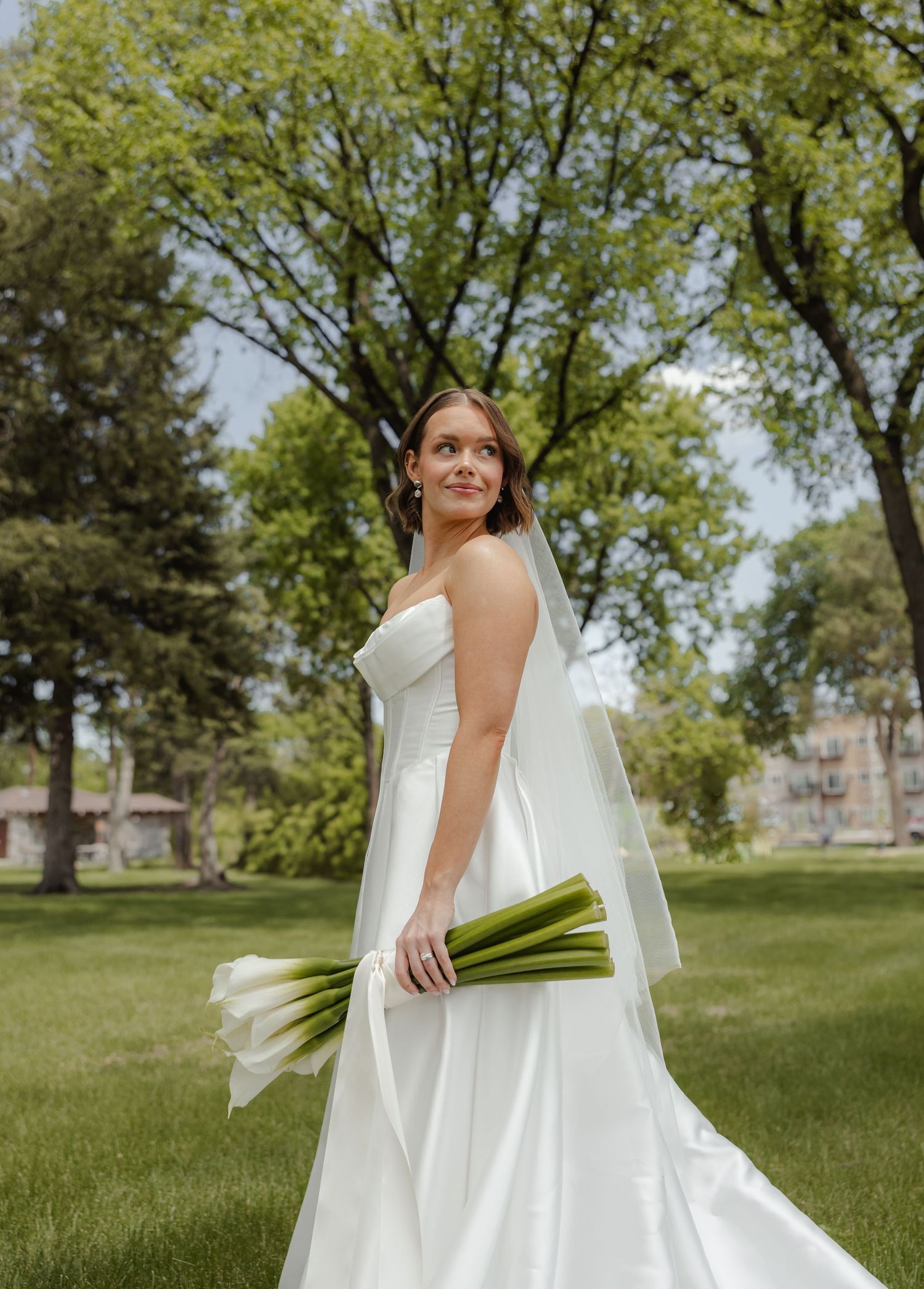 A bride in a flowing white wedding dress holds a bouquet while standing in a grassy park setting with trees.