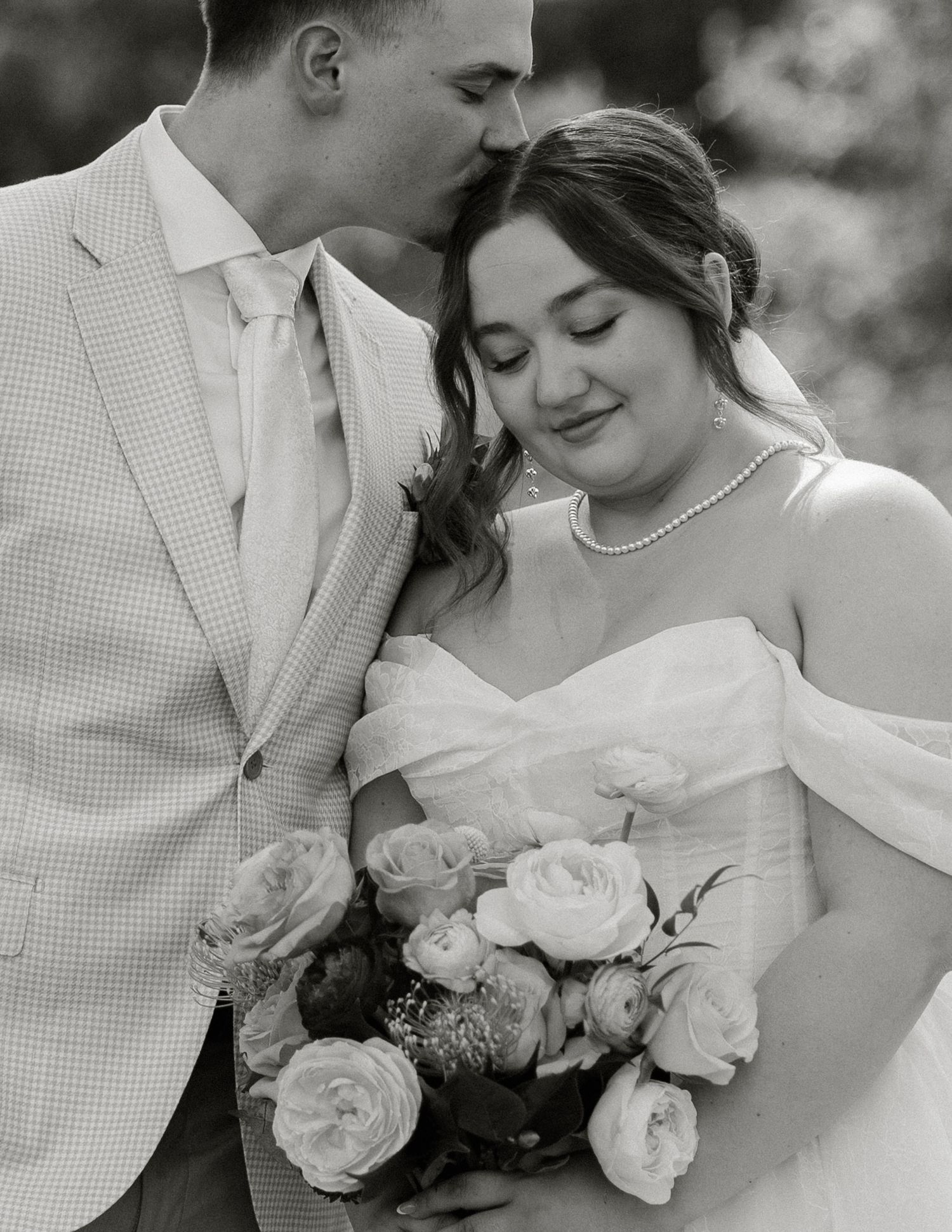 Black and white wedding photo showing tender moment between newlyweds with pearl necklace and rose bouquet.