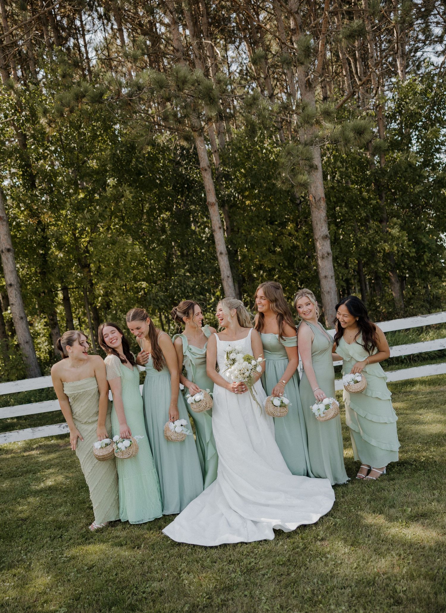 Bridesmaids in mint green dresses pose playfully with the bride on a grassy lawn near white fencing.