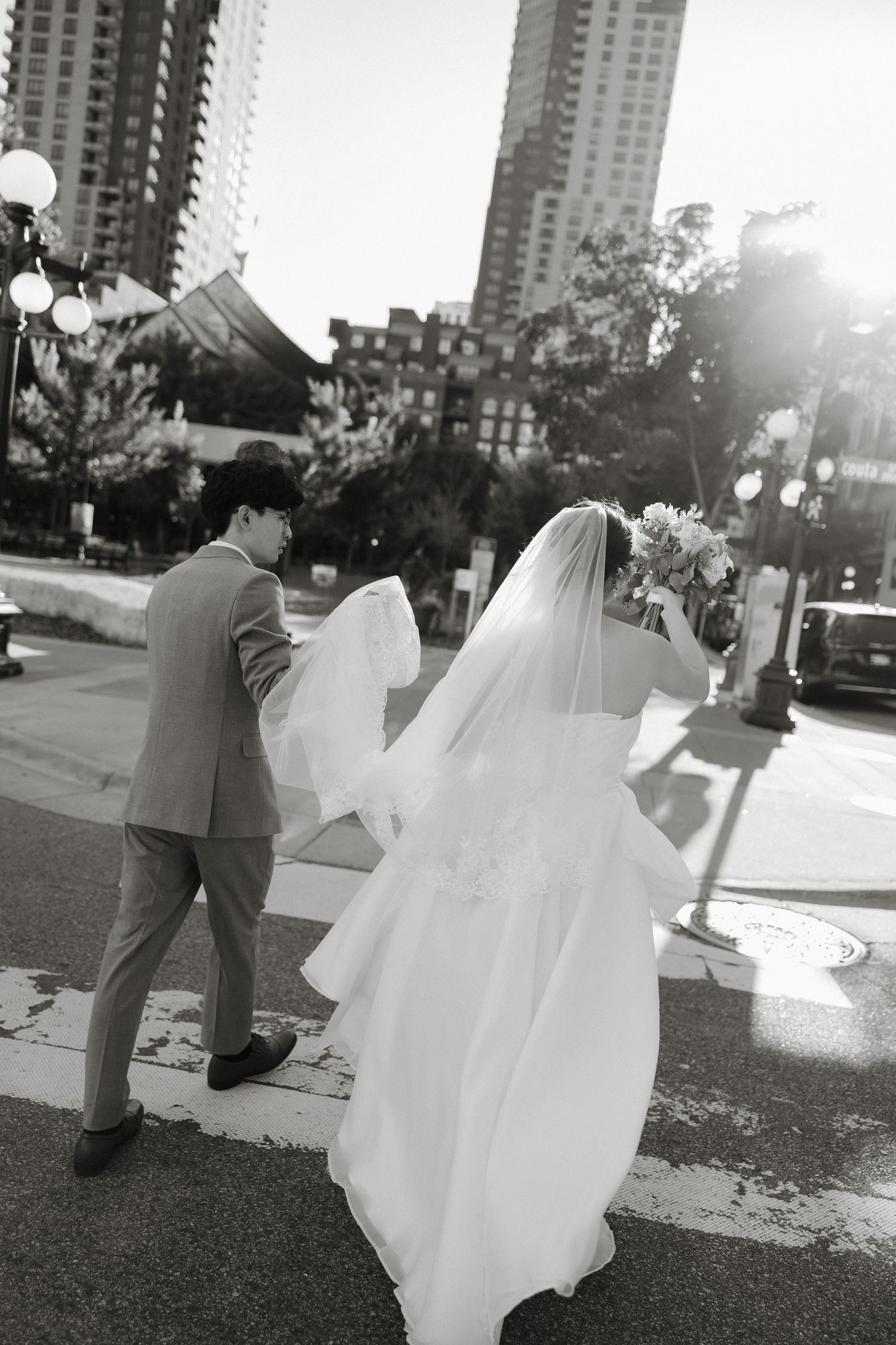 A black and white wedding photo of a couple walking down a city street with high-rise buildings in the background.