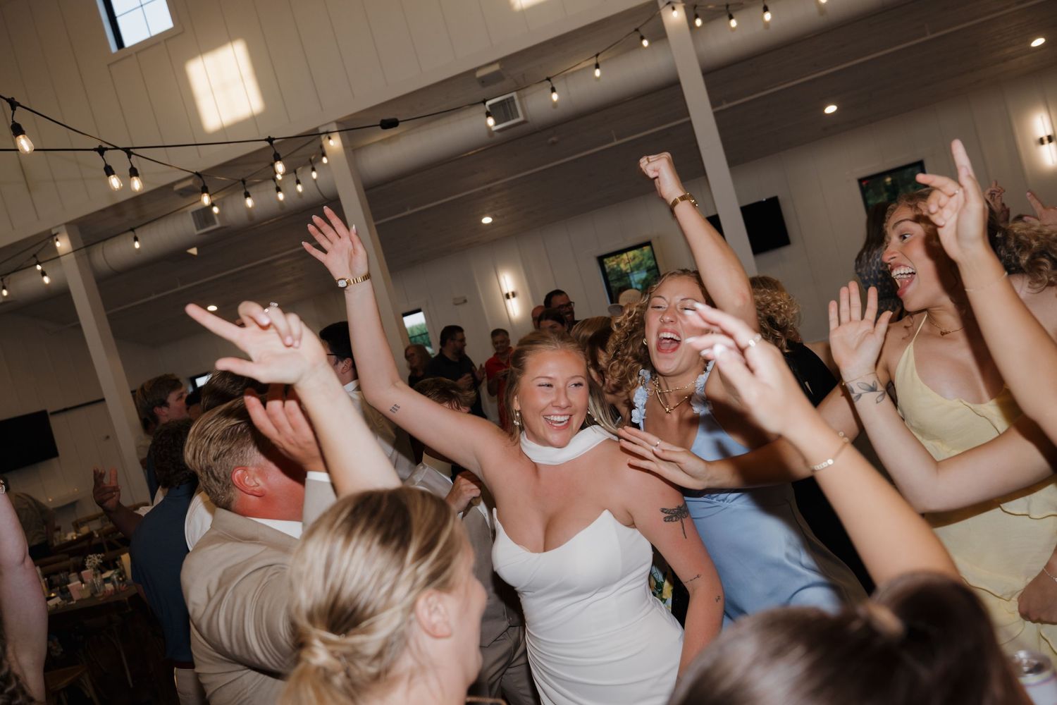 Wedding reception guests dance and celebrate with raised arms on a crowded dance floor under indoor lighting.