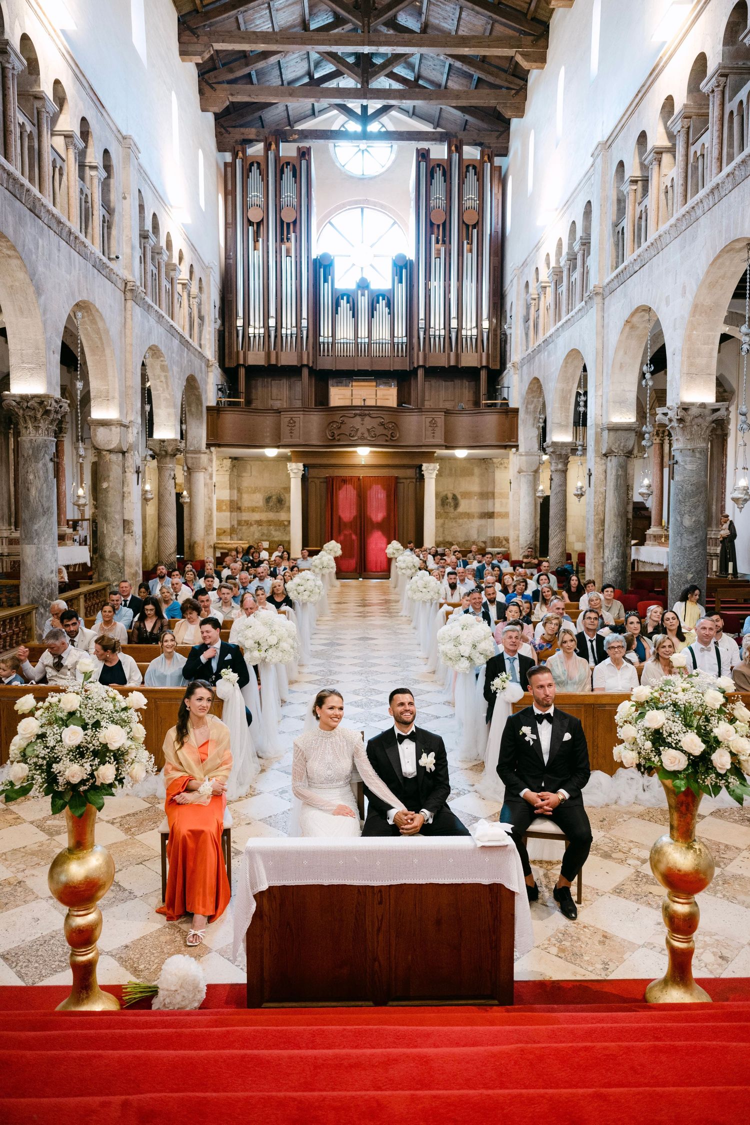 Bride and groom seated at altar during elegant church wedding ceremony with guests, floral arrangements, and pipe organ backdrop.