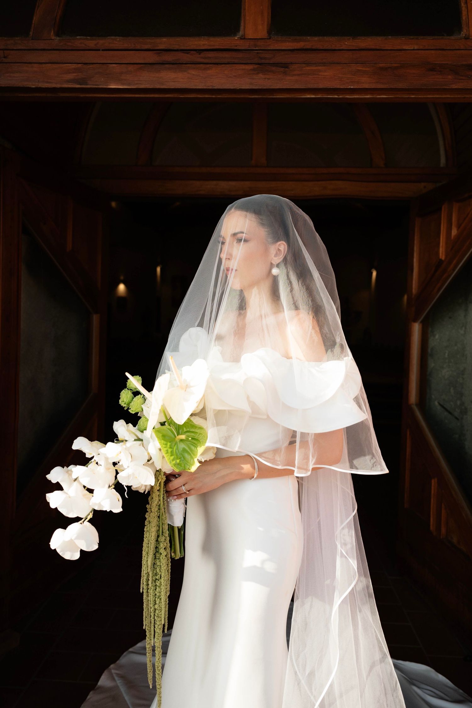 Elegant bride in white off-shoulder gown and veil holding white orchid bouquet in a dark wooden venue.