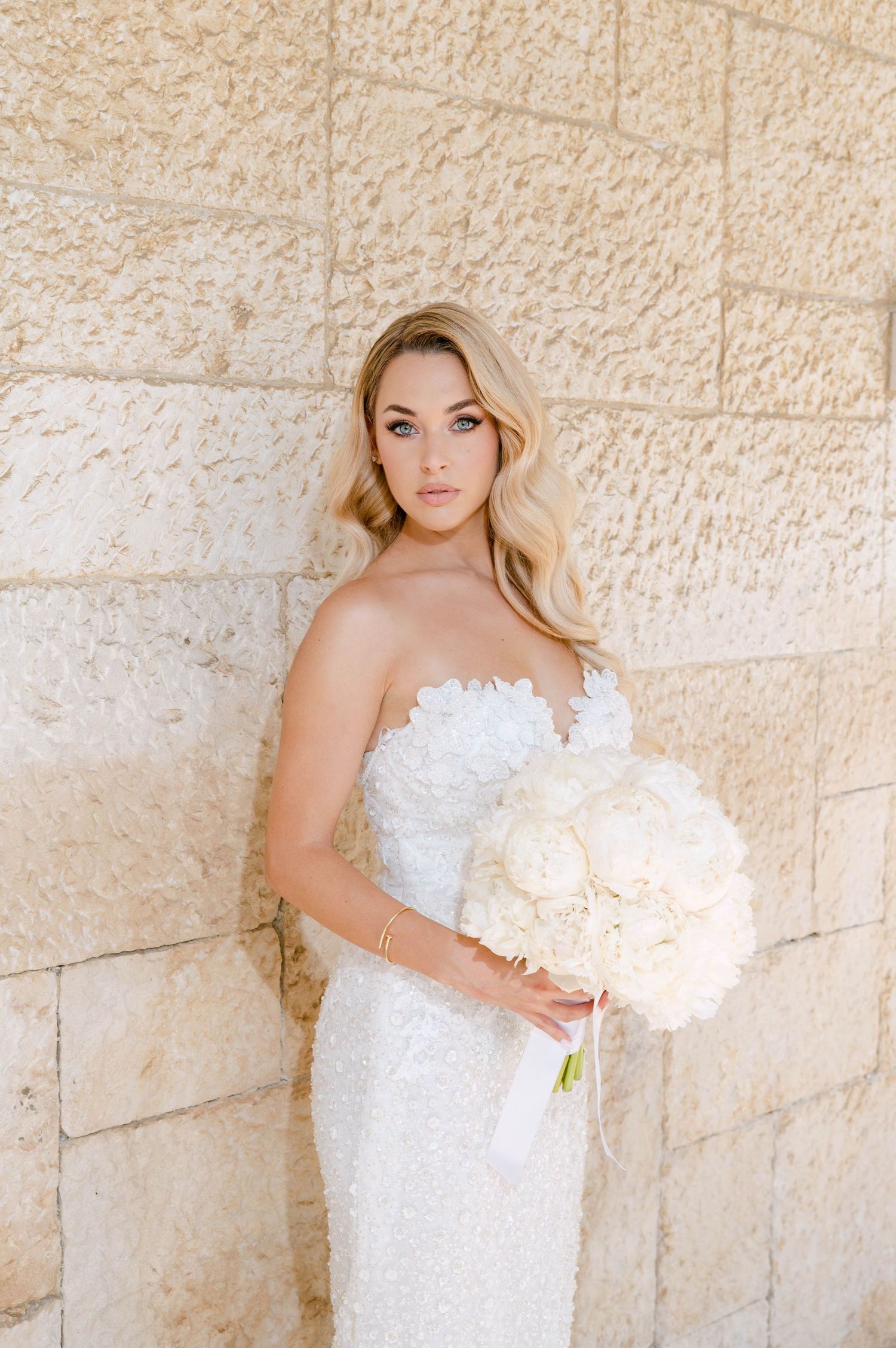 A bride in a white strapless wedding dress poses against a textured stone wall holding a light-colored bouquet.