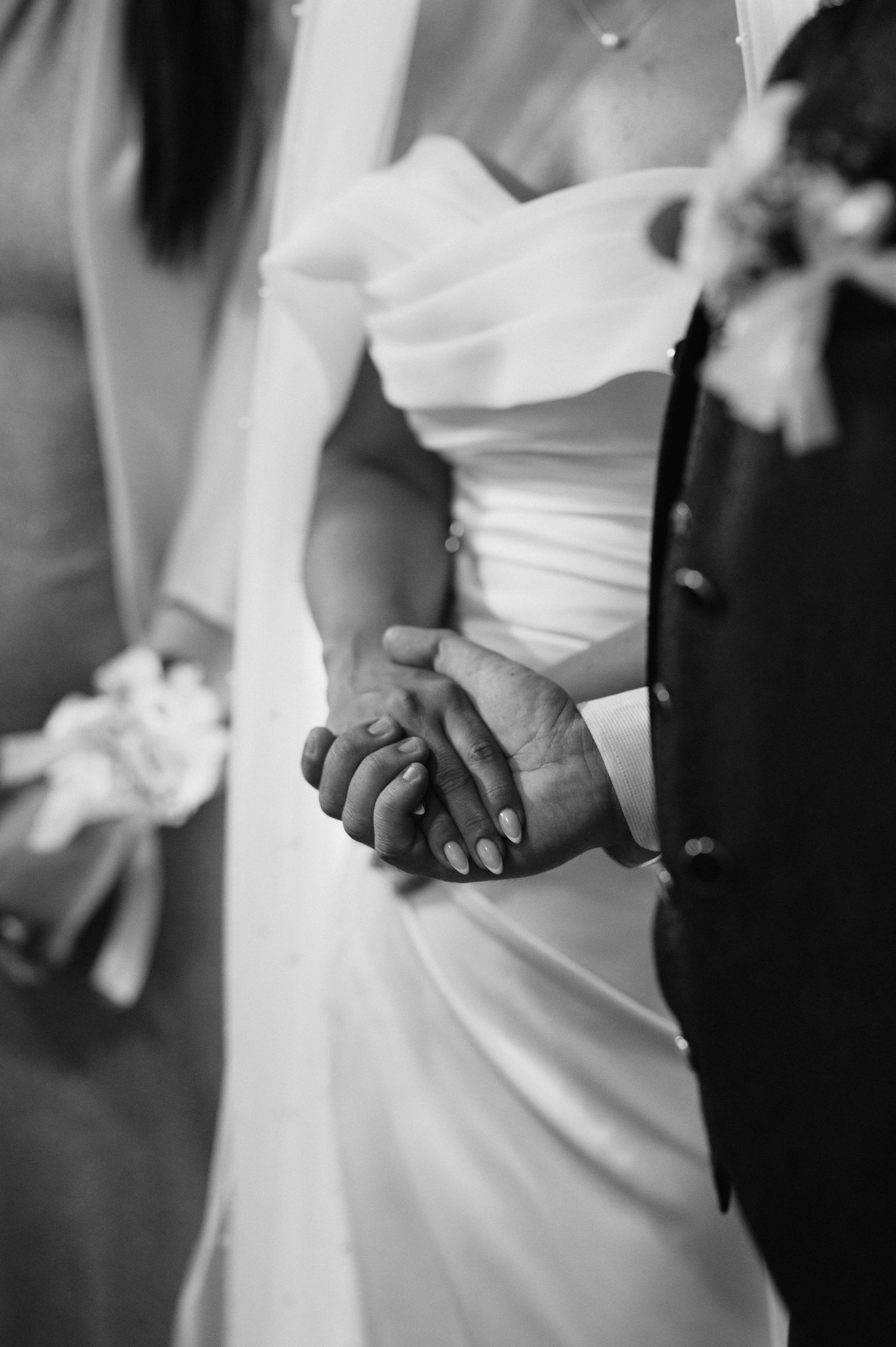 A black and white photo captures an intimate moment of newlyweds holding hands, with the bride in a white wedding dress.