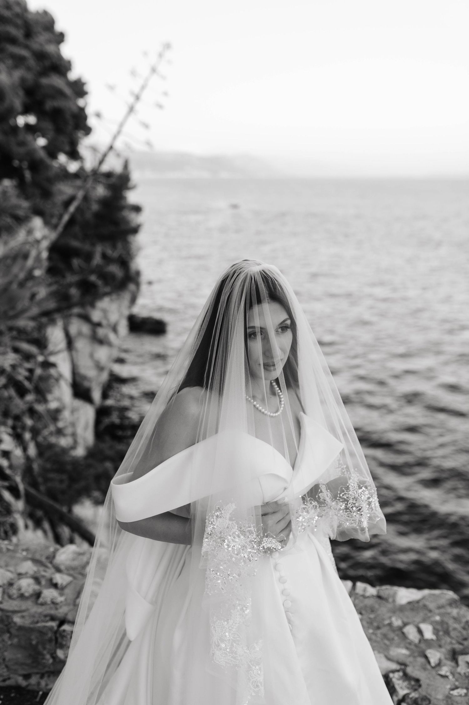 A scenic black and white bridal portrait by the ocean shore featuring an elegant lace wedding dress and flowing veil.