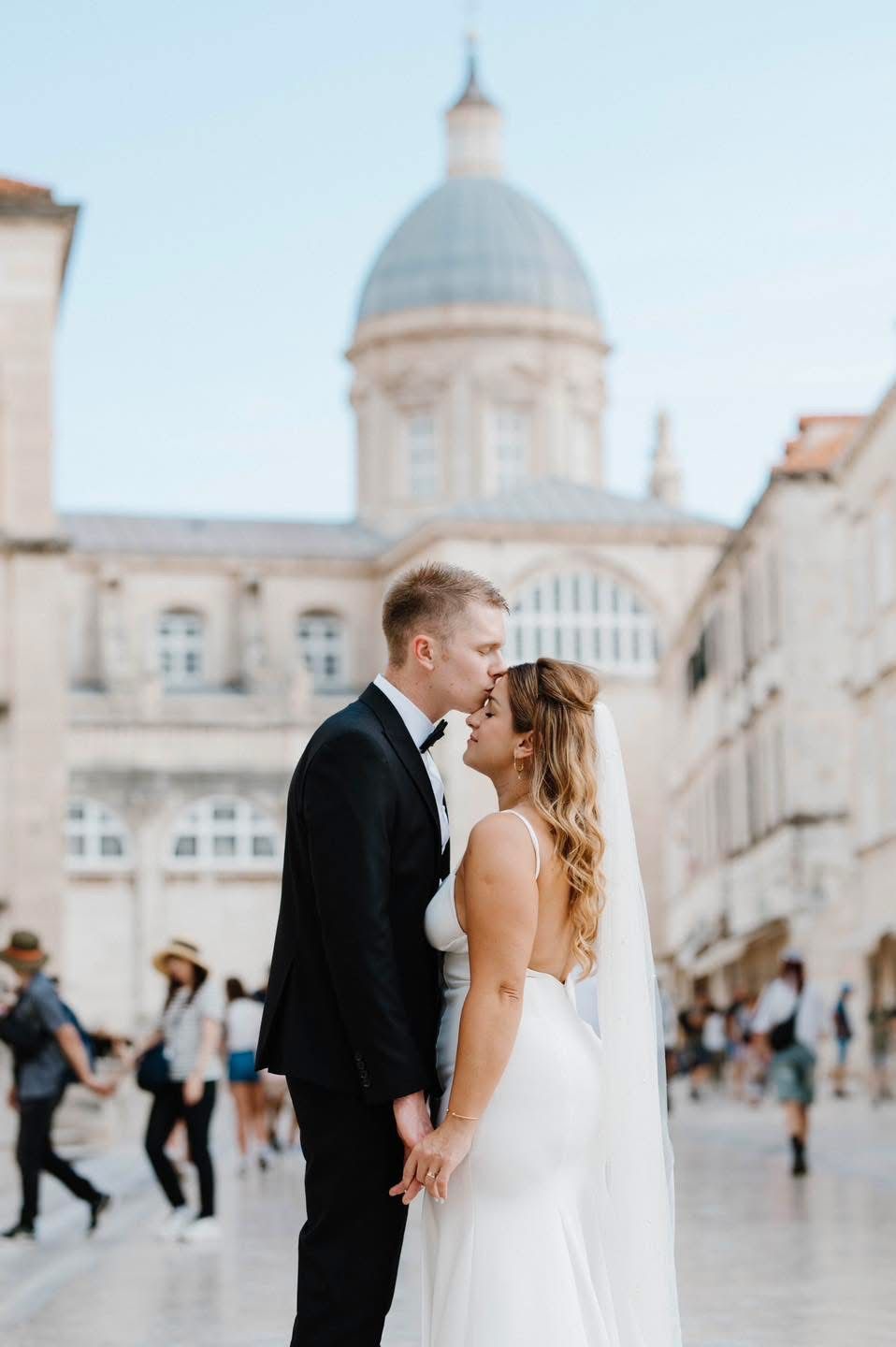 Wedding couple shares intimate moment in front of historic domed building in European city square.