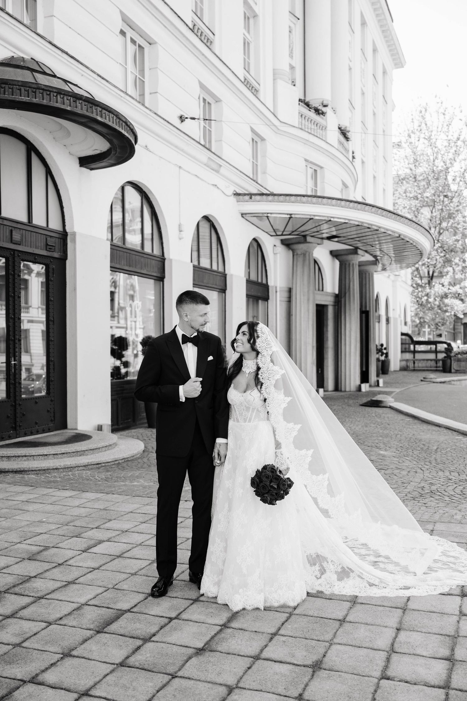 Black and white wedding photo of bride in lace gown with veil and groom in tuxedo walking outside elegant building.