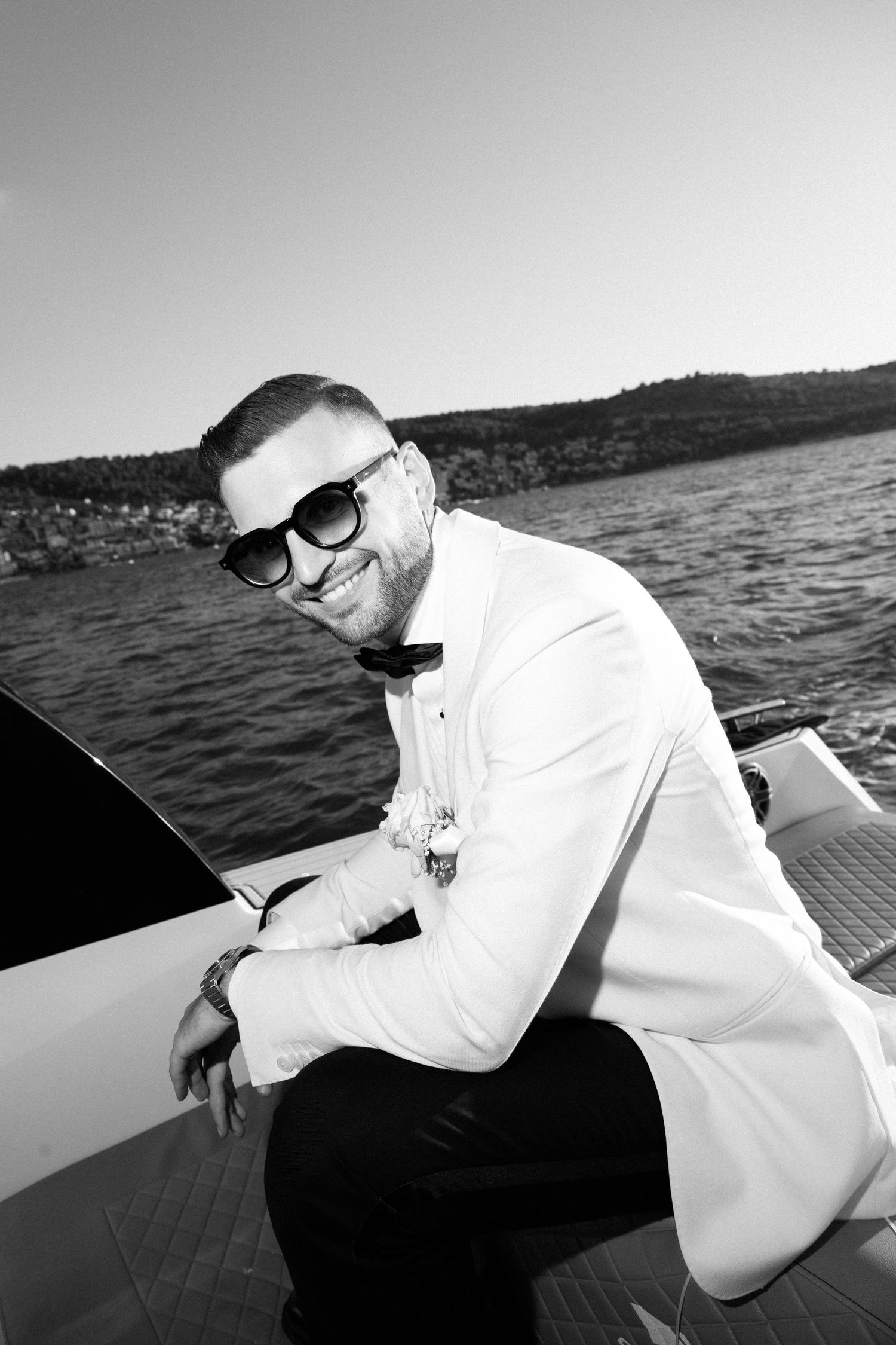 Smiling man in white tuxedo jacket and sunglasses sitting on a luxury yacht with ocean backdrop, black and white photo.