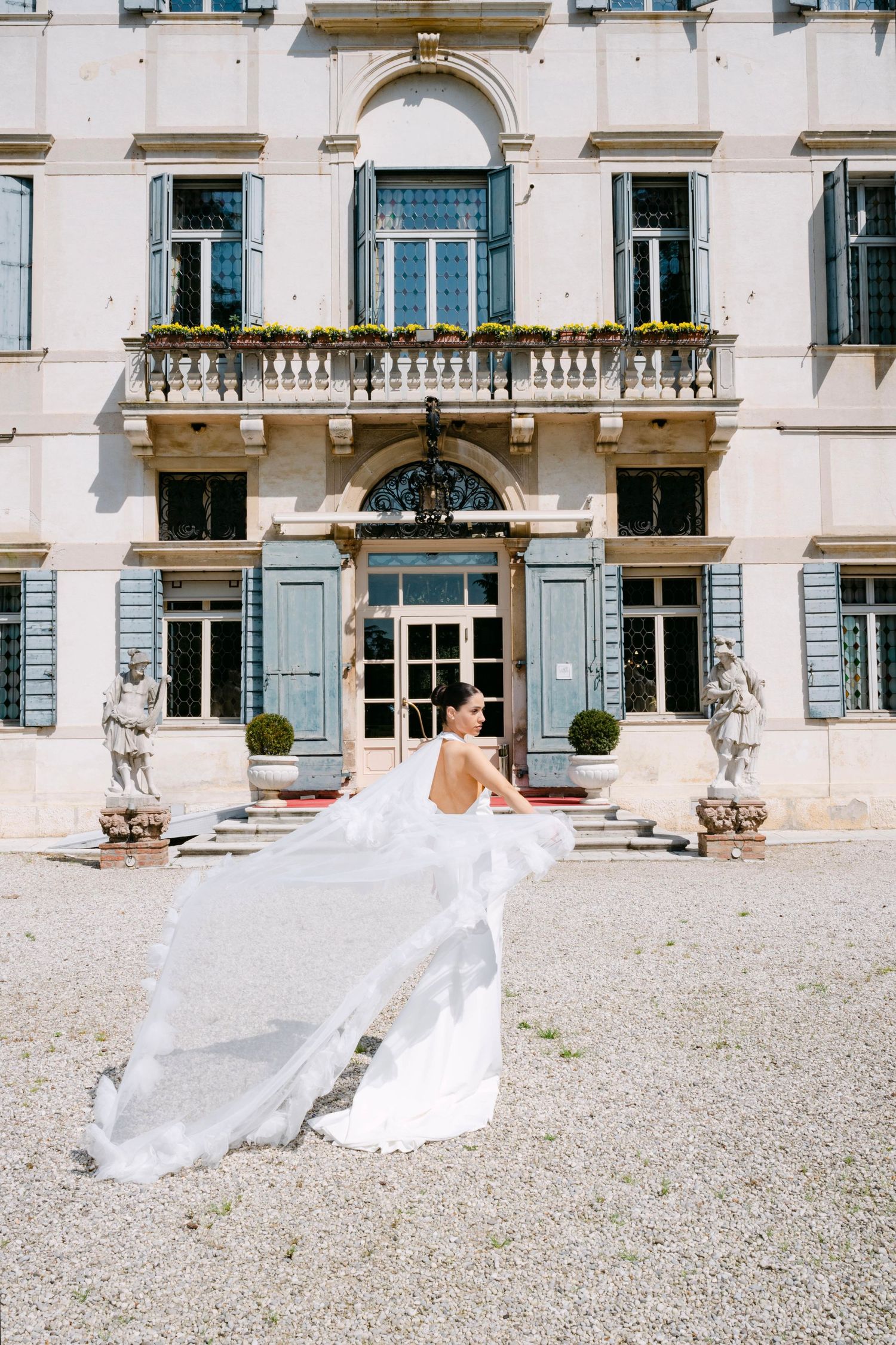 Elegant white flowing dress billows in the breeze in front of a grand European-style villa with ornate architectural details.