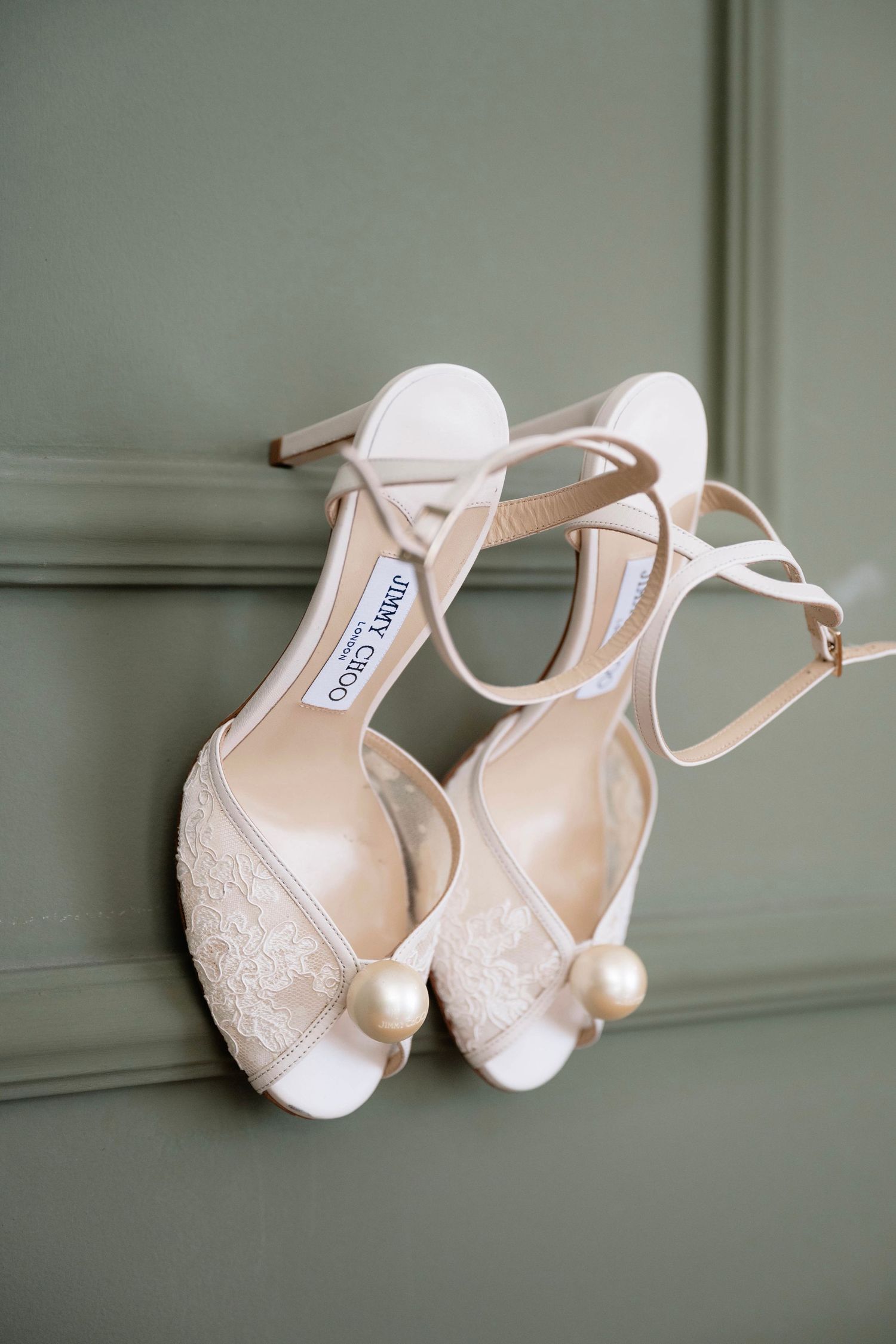Jimmy Choo white lace bridal heels with pearl embellishments and ankle straps displayed against a sage green wall.