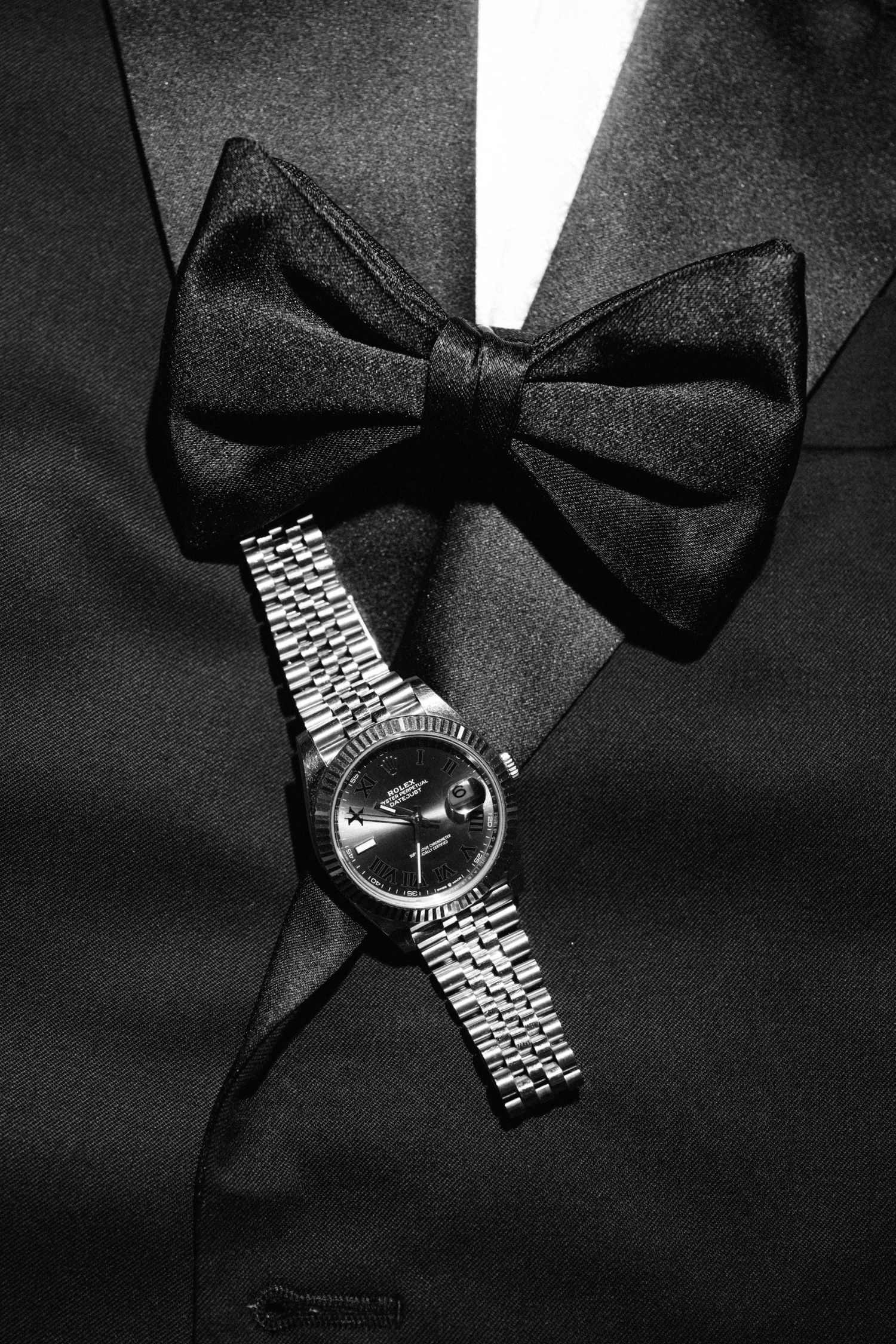 Black and white photo of a Rolex watch resting on a tuxedo jacket with a satin bow tie.