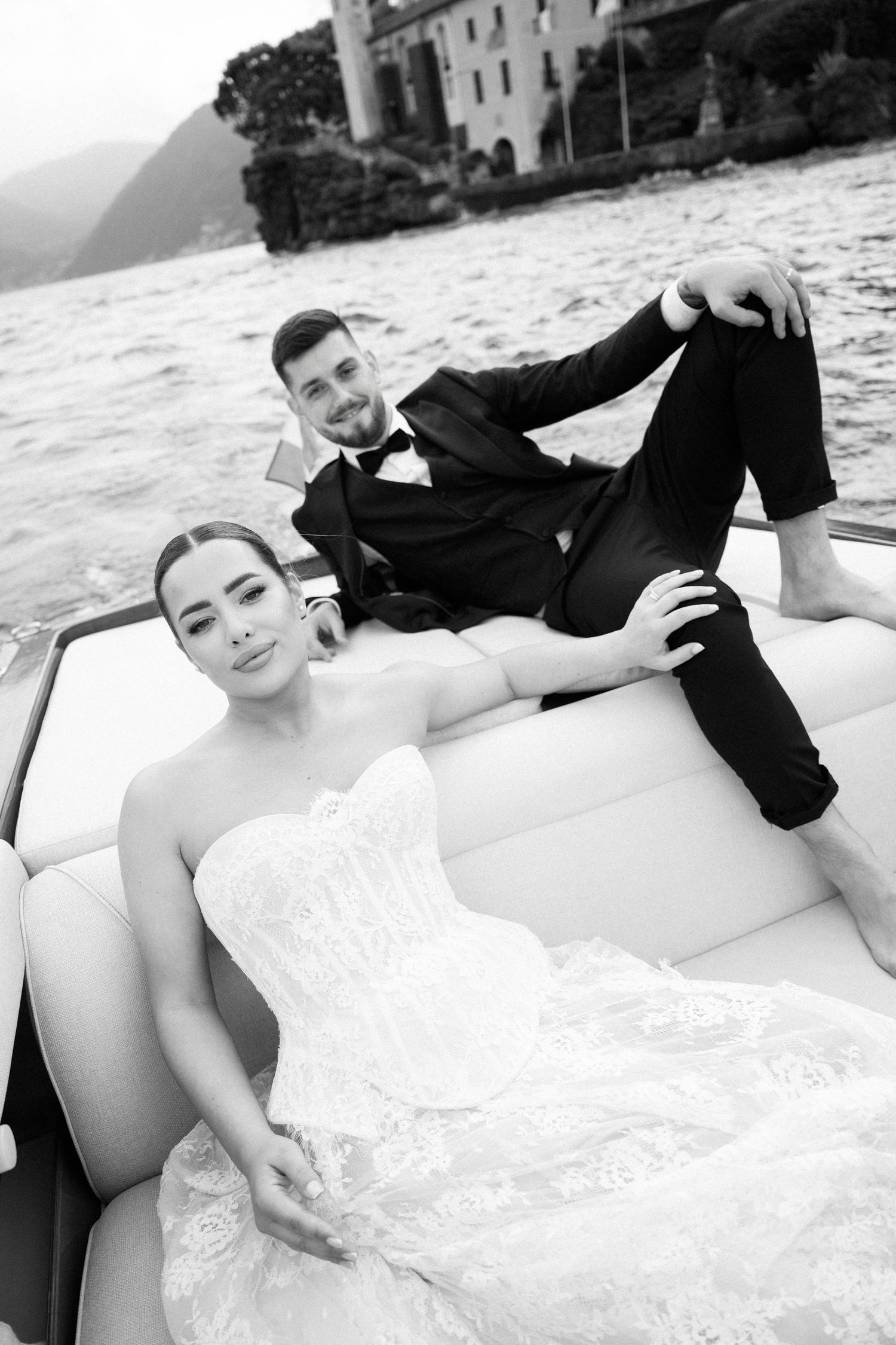 Black and white photo of bride and groom relaxing on a luxury boat with a lakeside villa in the background.