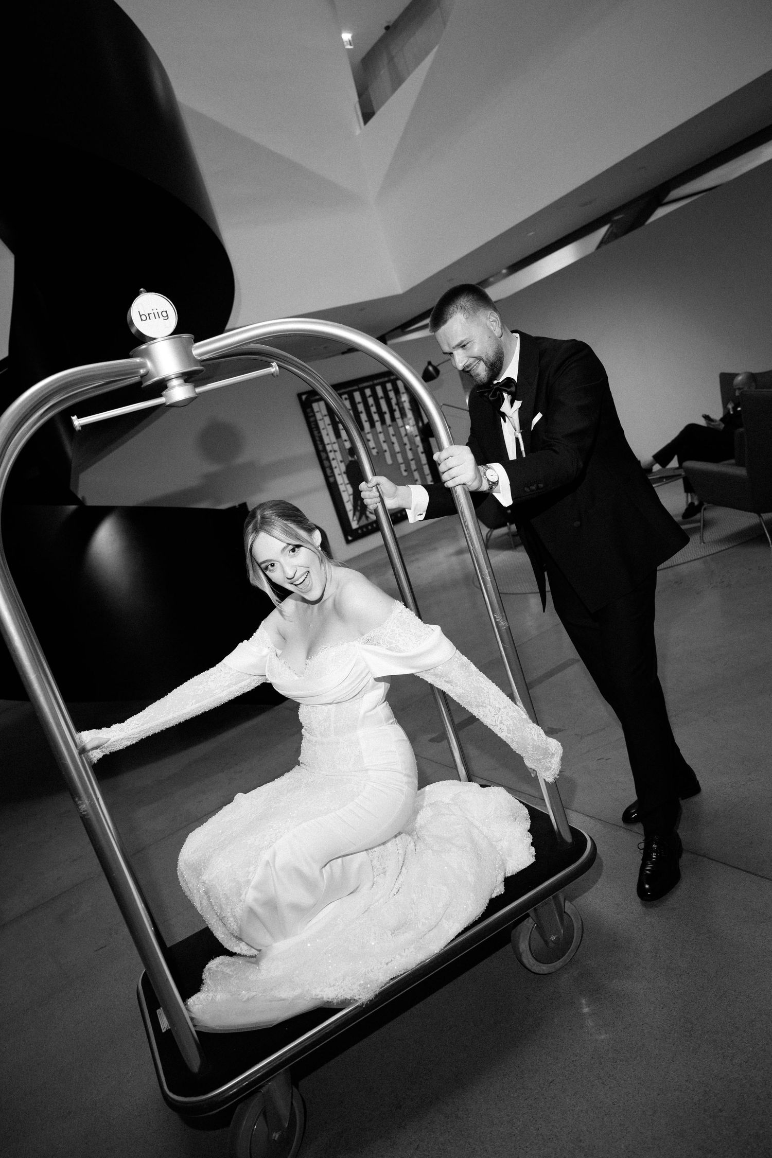 Bride in white gown rides a hotel luggage cart pushed by groom in tuxedo, laughing in black and white photo.